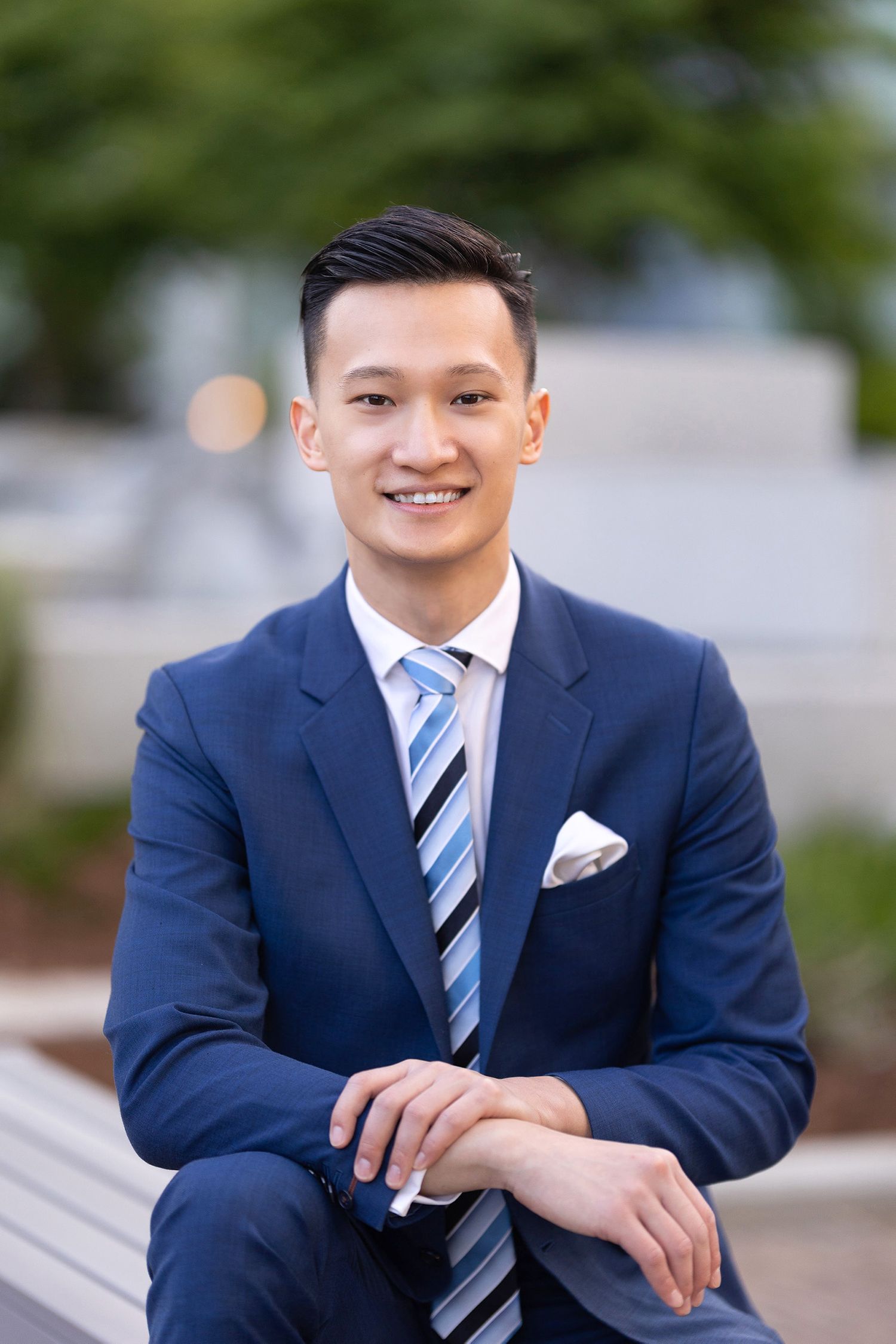 Smiling young Asian man in a blue suit sitting outdoors on a bench with green bokeh background.