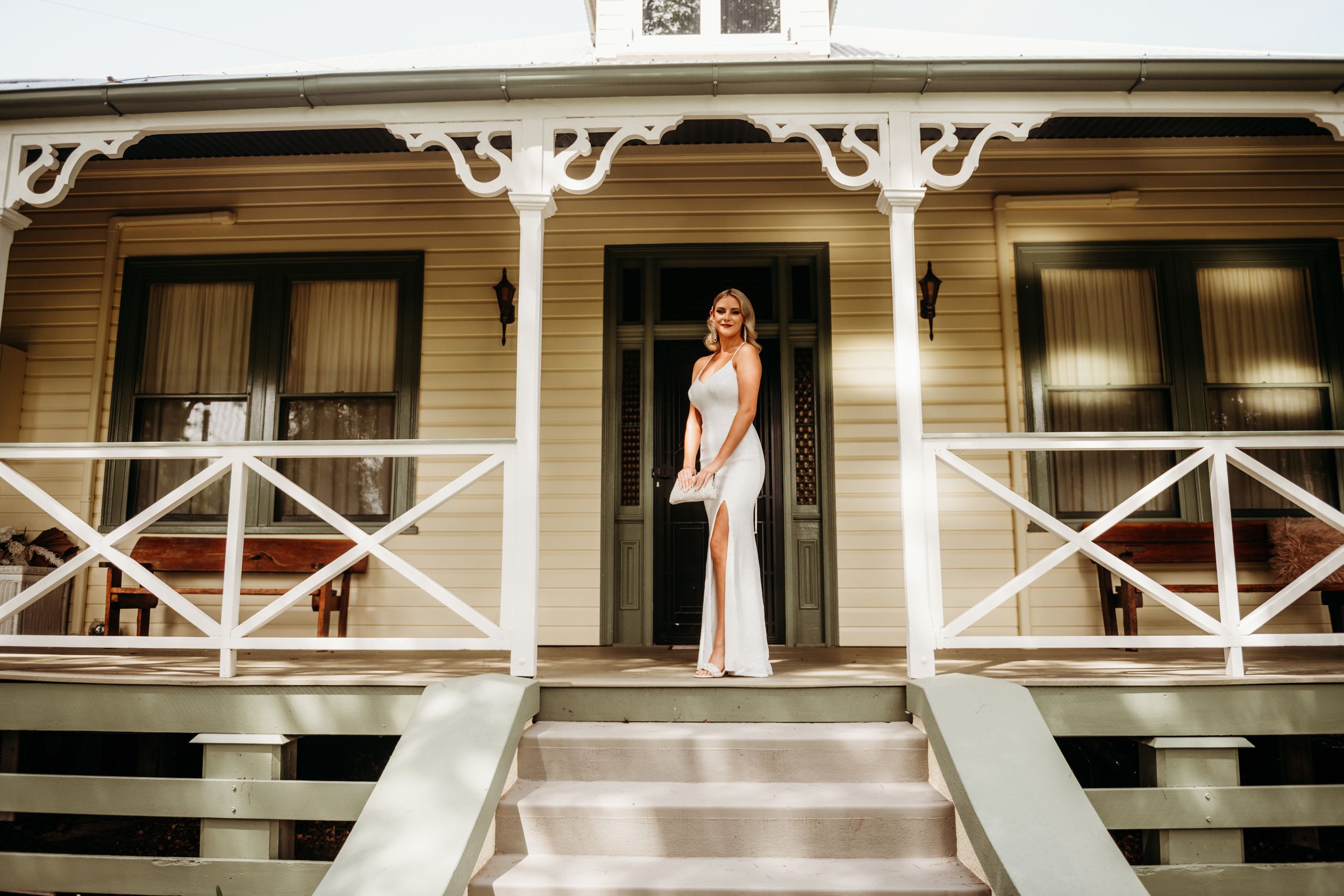 A person in a white dress poses on the steps of a classic Victorian-style porch with ornate trim work.