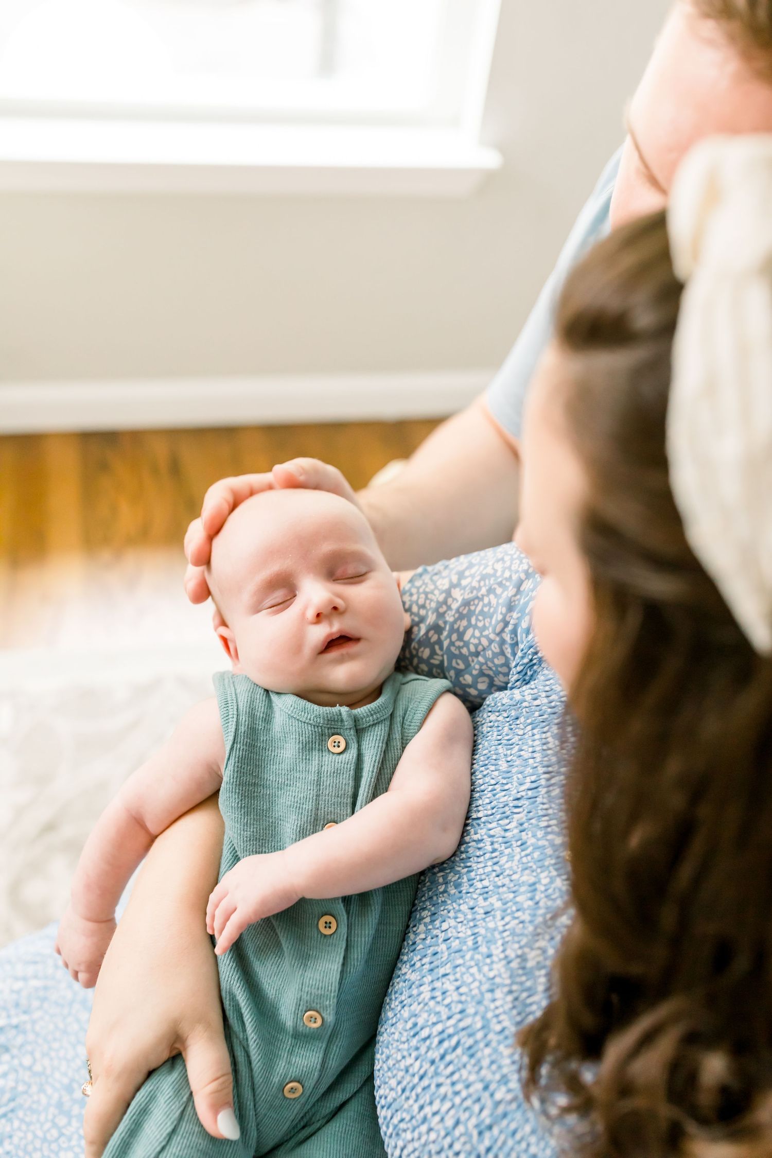 A sleeping infant in a teal romper being held gently against soft white bedding.