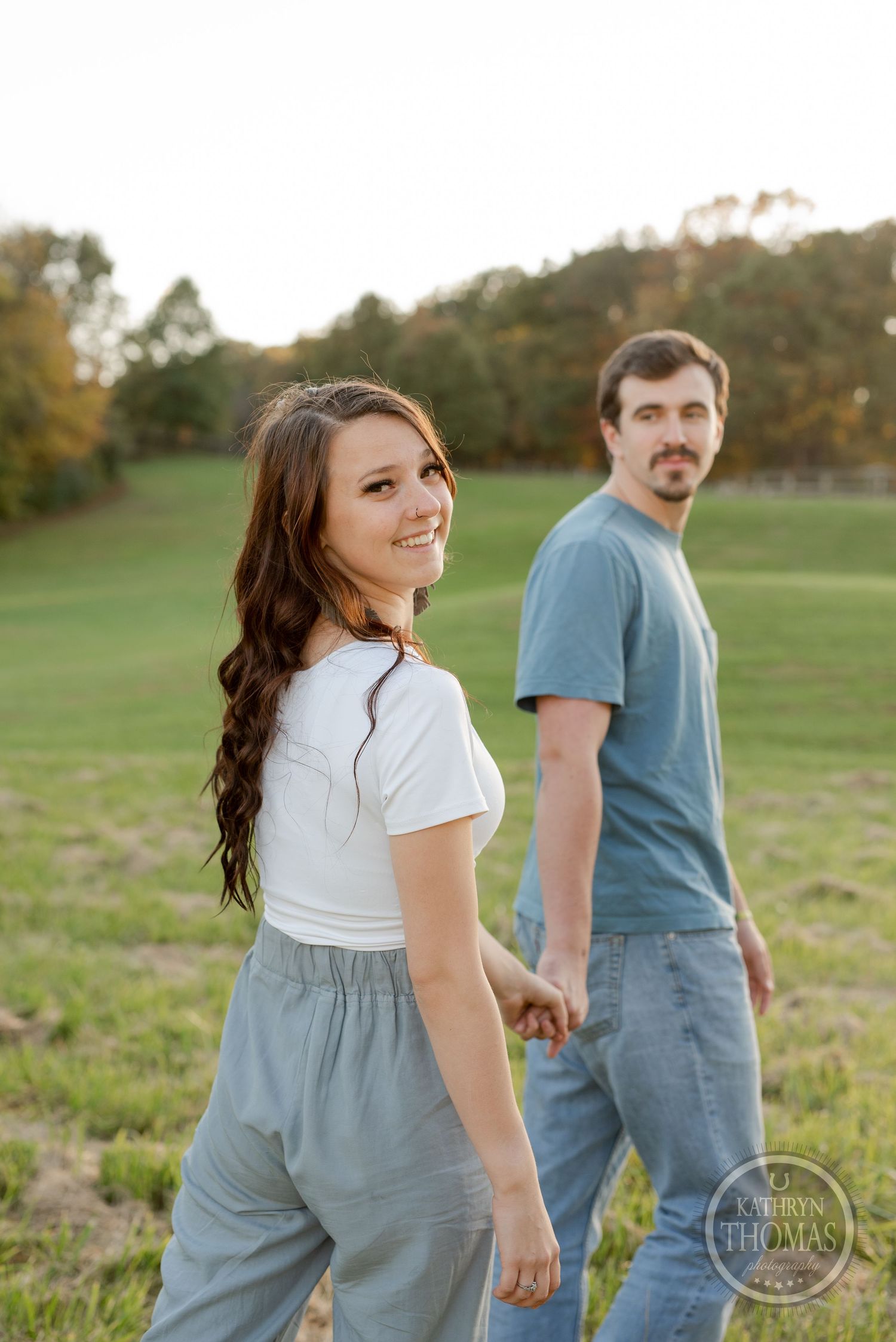 A couple looks back at the camera while walking hand in hand through a grassy field.