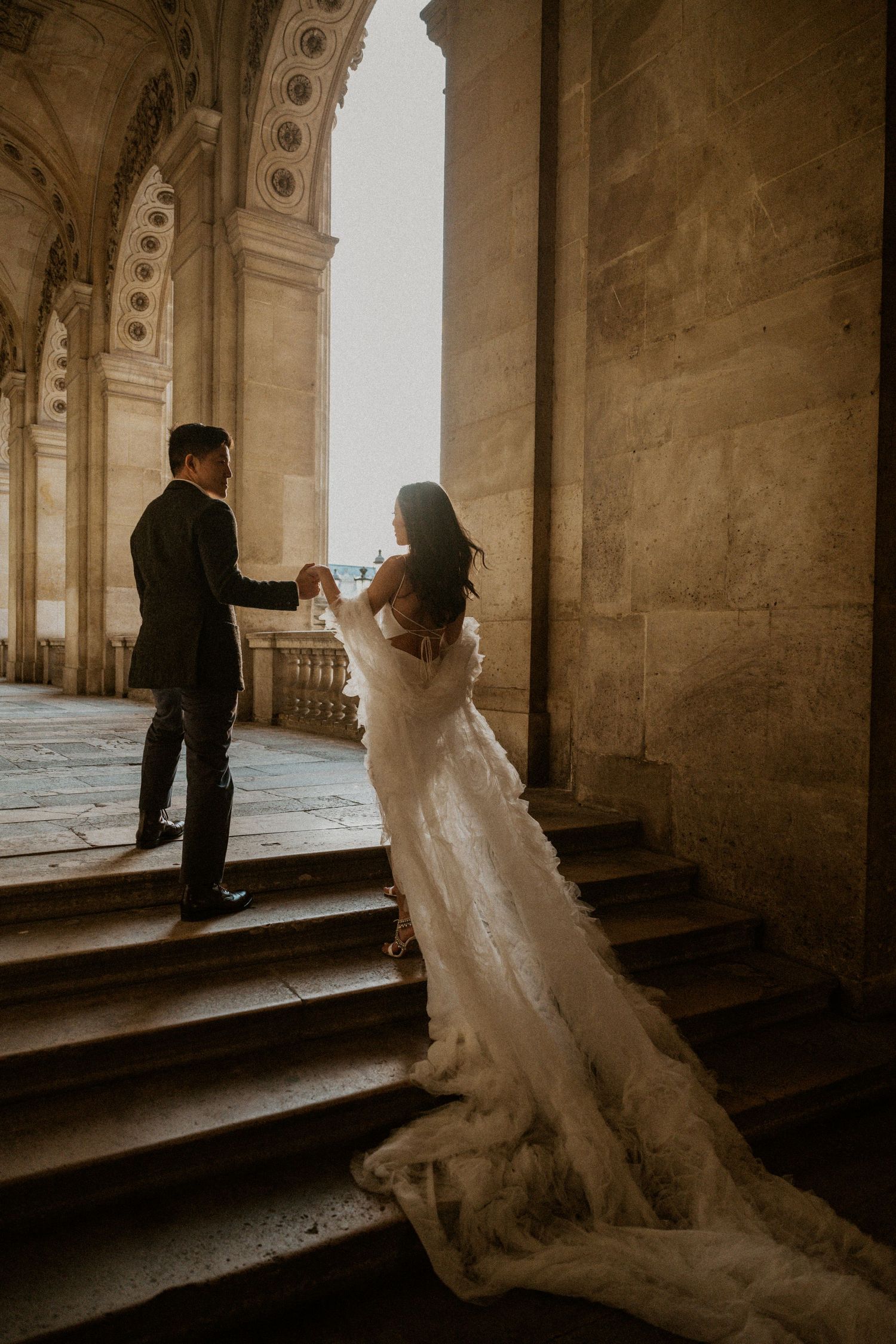 Couple in wedding attire poses on grand stone staircase in warm sunlight.