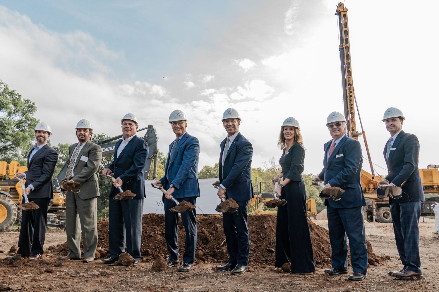 Group of people in business attire participating in a groundbreaking ceremony with shovels at a construction site.