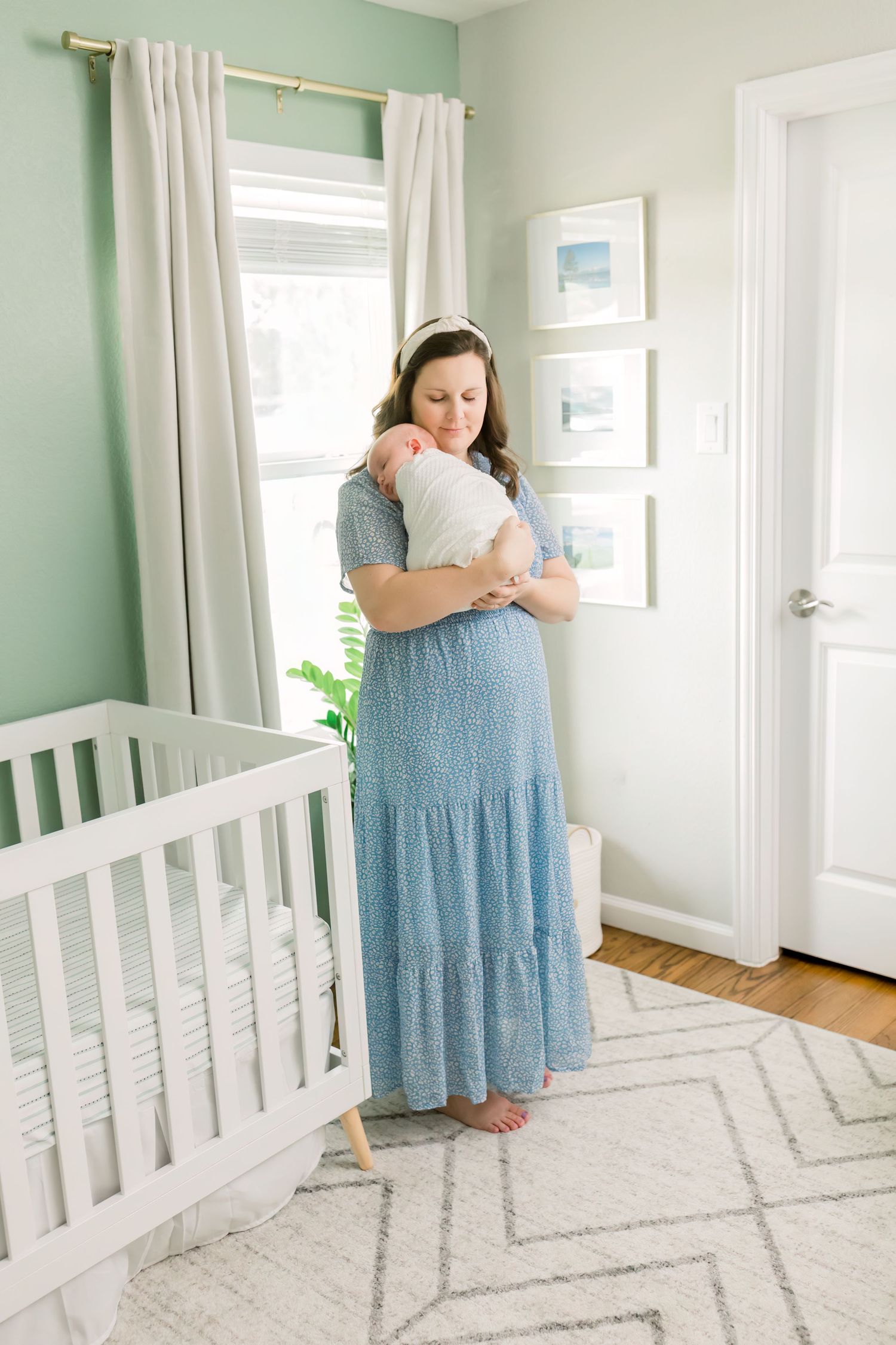 Woman in flowing blue dress holding swaddled infant in mint green nursery.