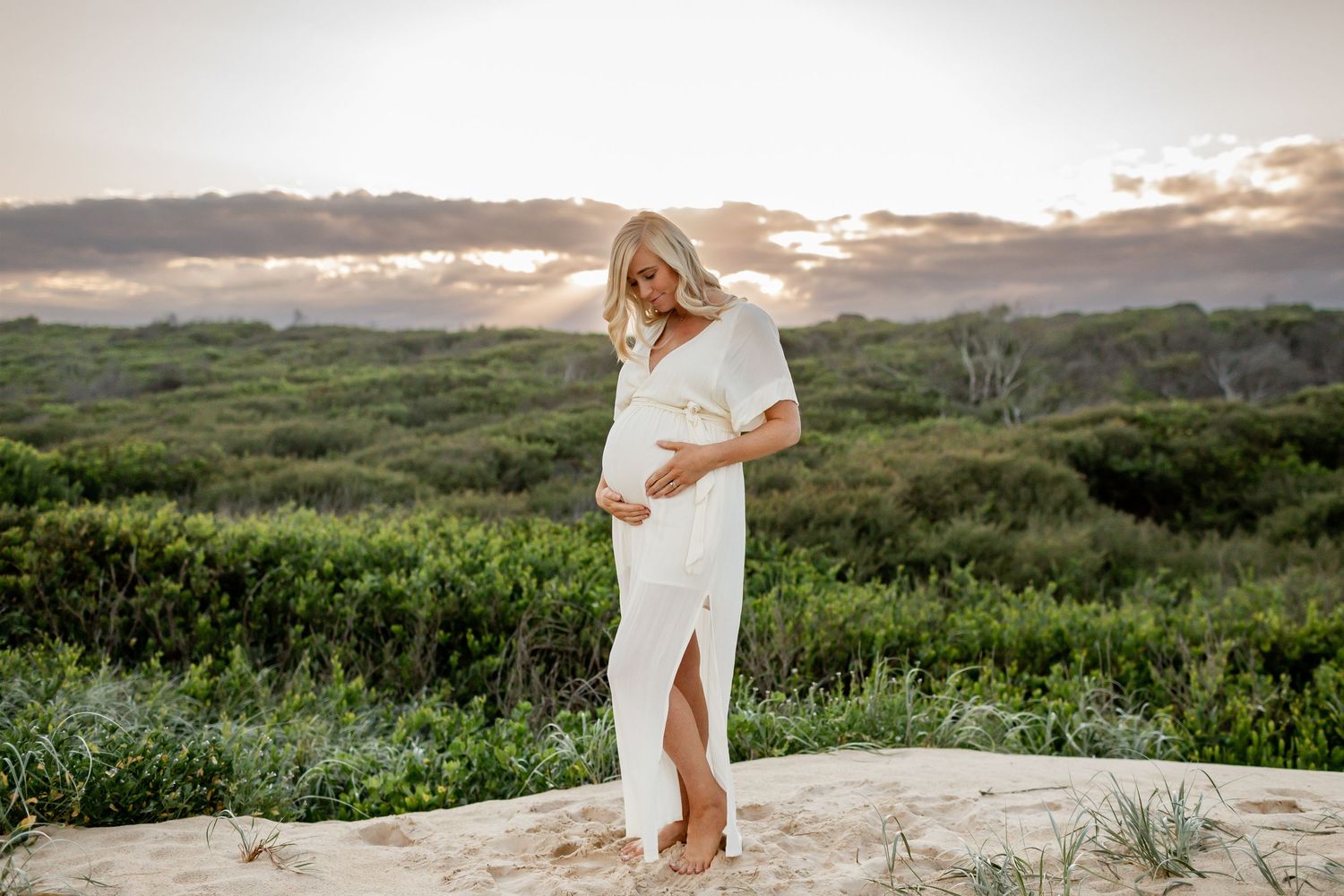 Pregnant woman in white dress standing on rocky outcrop during sunset.