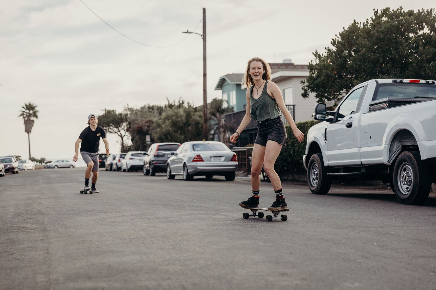 People skateboarding down a street lined with parked cars on a cloudy day.