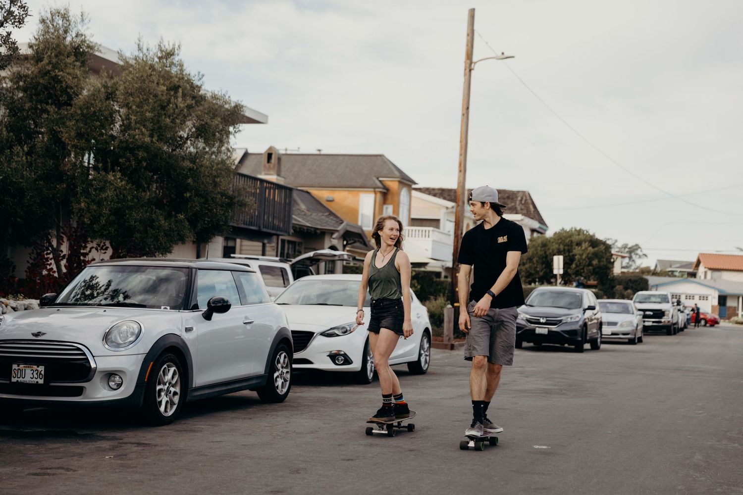 Two people skateboarding together on a residential street lined with parked cars.