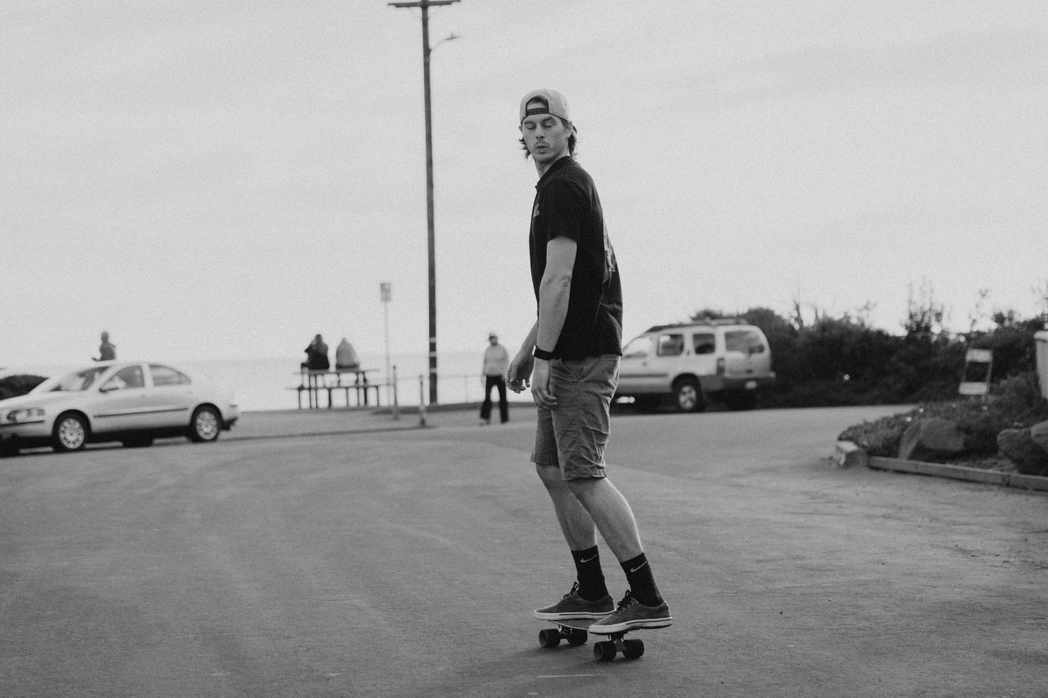 Black and white photo of someone skateboarding in a parking lot with cars in the background.
