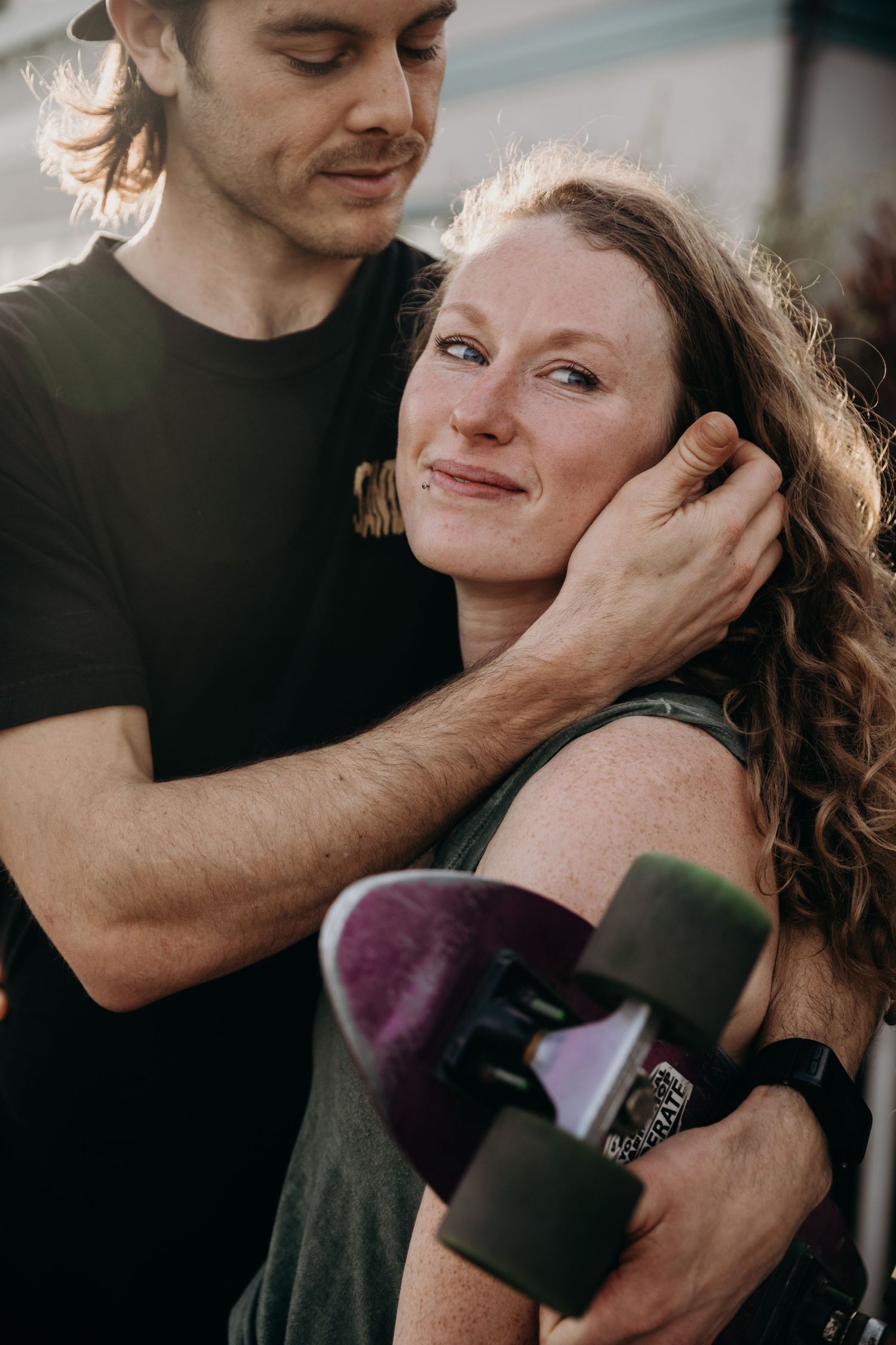 A couple shares an intimate moment outdoors while holding a skateboard in casual attire.