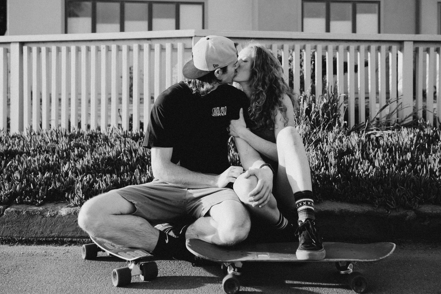 A romantic black and white moment captured of a couple sitting on skateboards near a white fence.