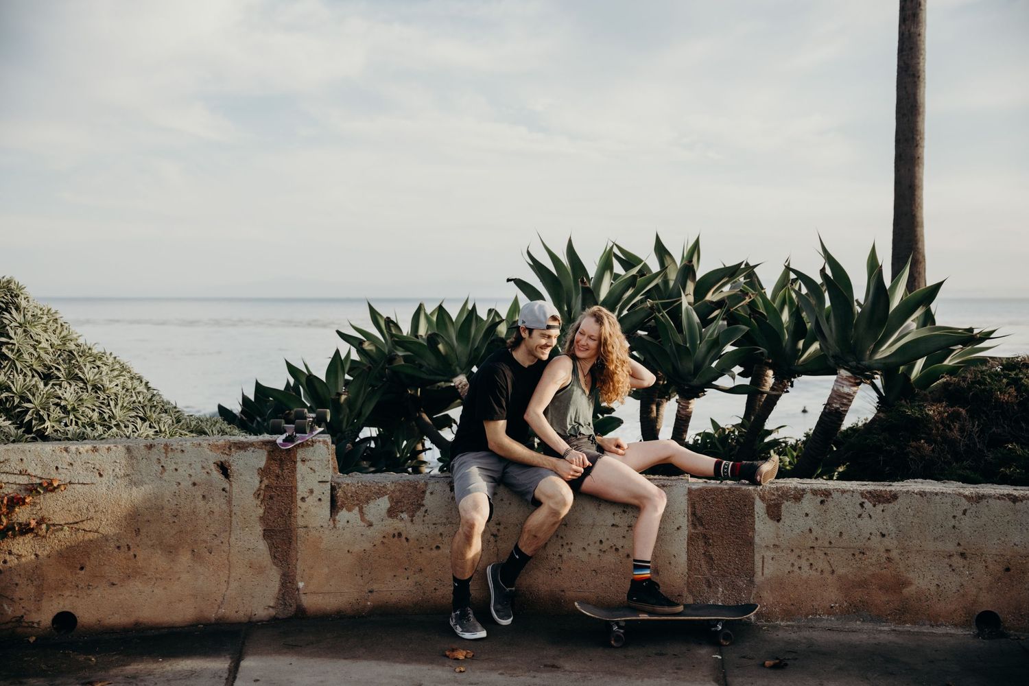 Two people sitting on a concrete wall with ocean and plants in the background.