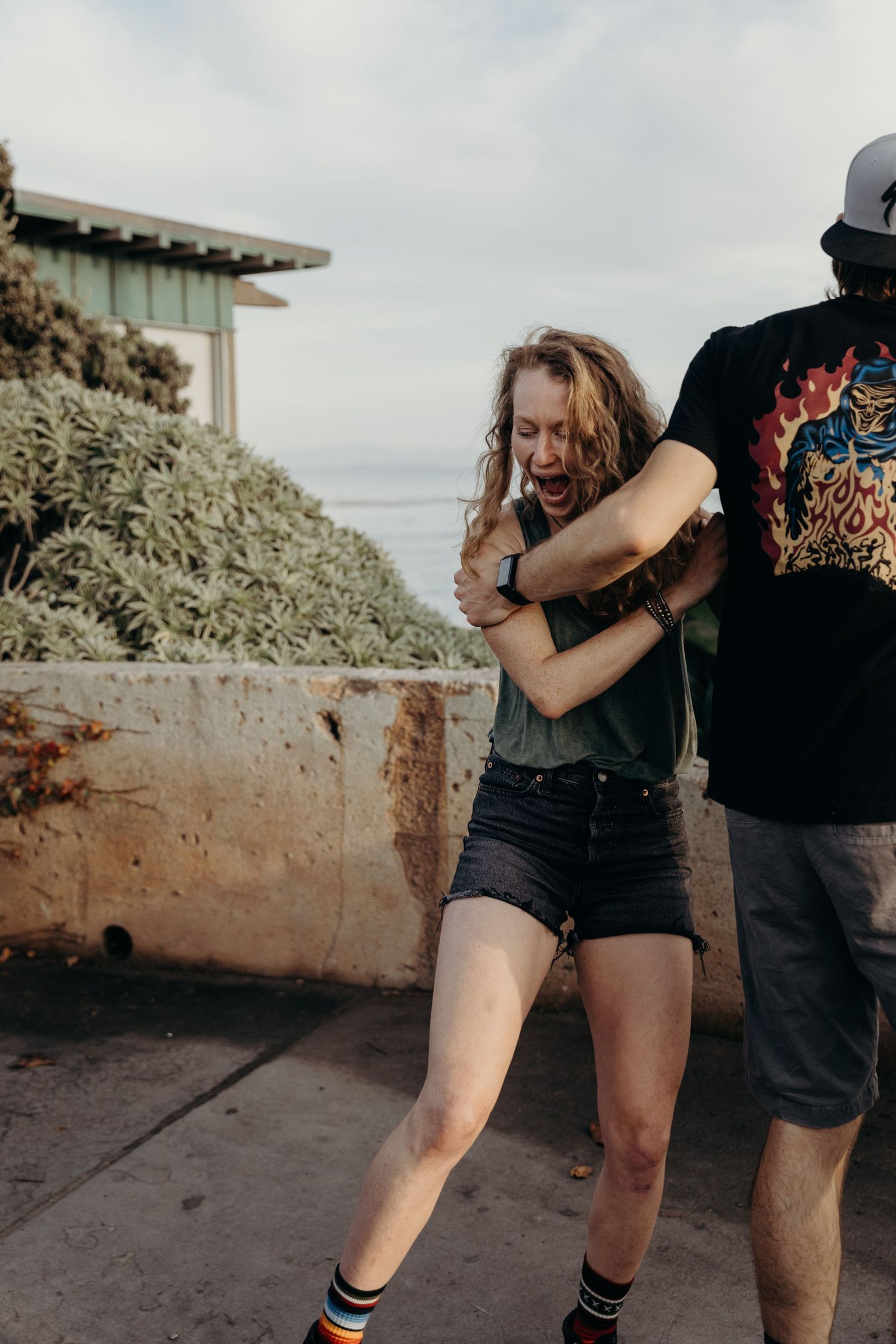 A playful moment between two people near a concrete wall with coastal vegetation.