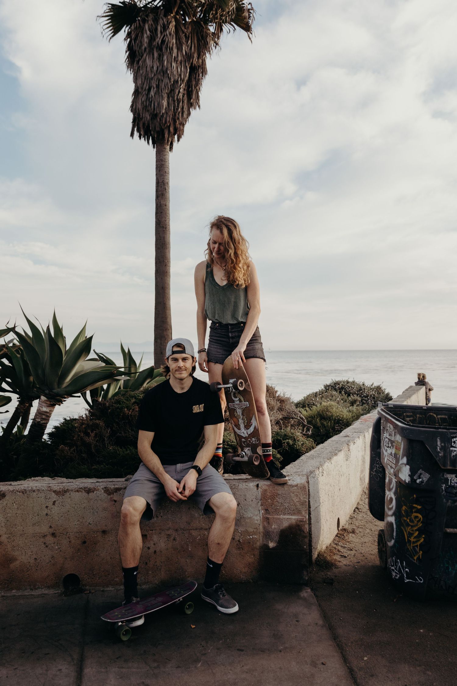Young couple with skateboards posing by palm trees and ocean on sunny coastal wall.