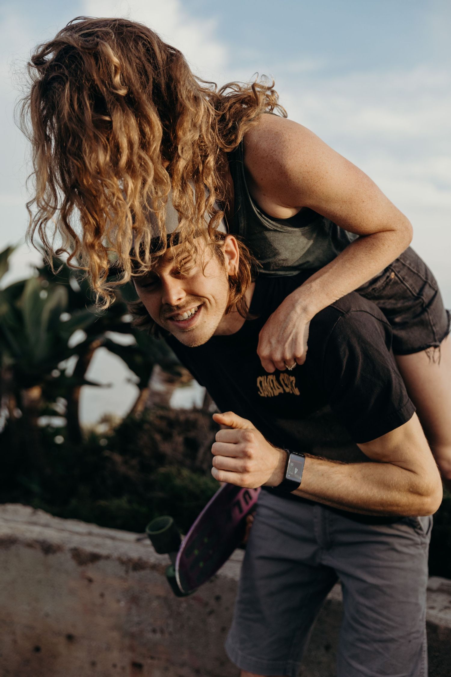 Playful couple sharing a joyful moment outdoors with skateboard near palm trees.