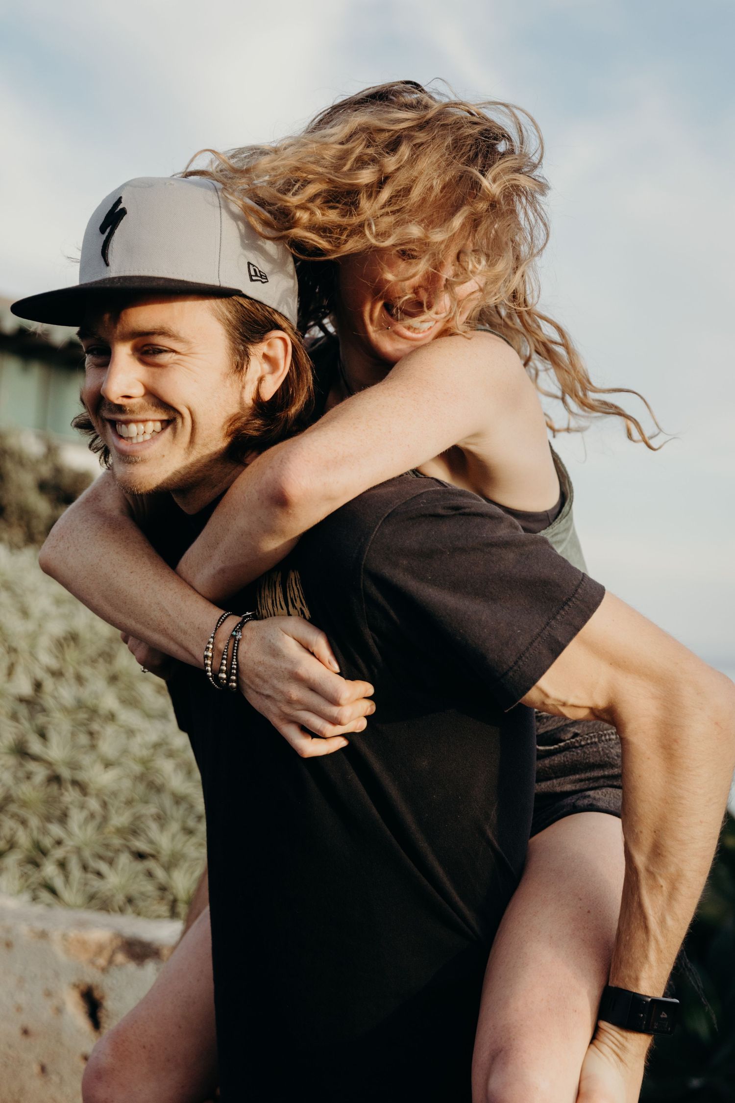Couple enjoying a piggyback ride outdoors on a sunny day near the coast.