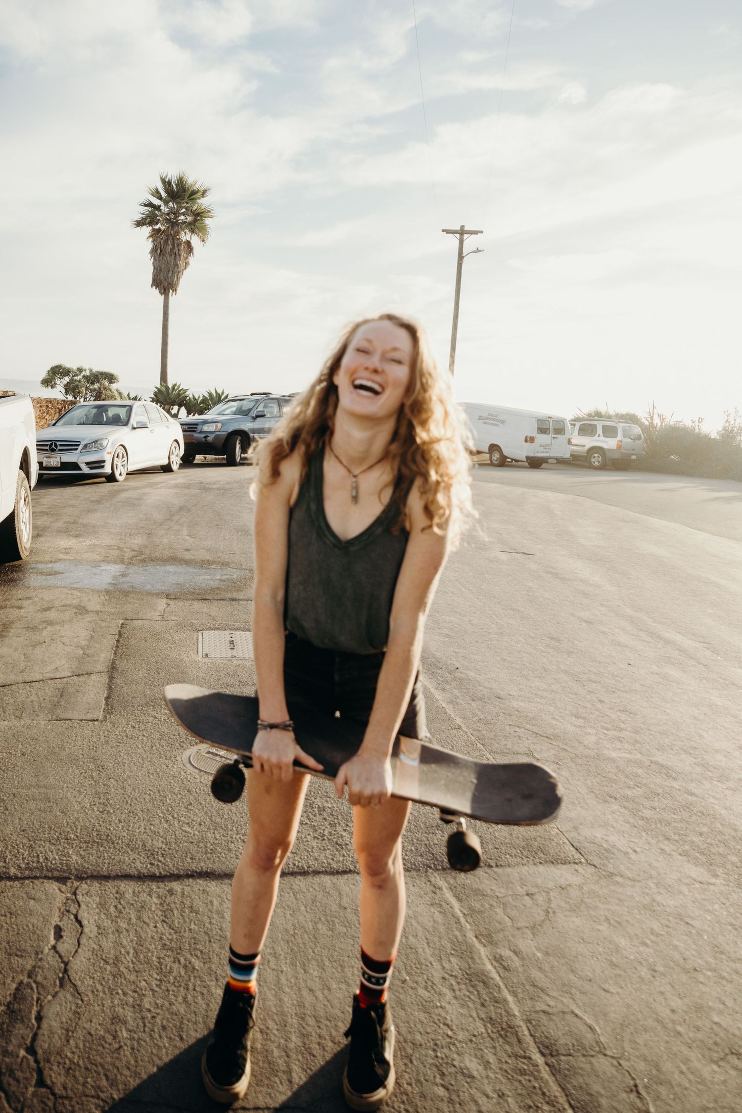 Person with long wavy hair laughing while holding skateboard in sunny parking lot.