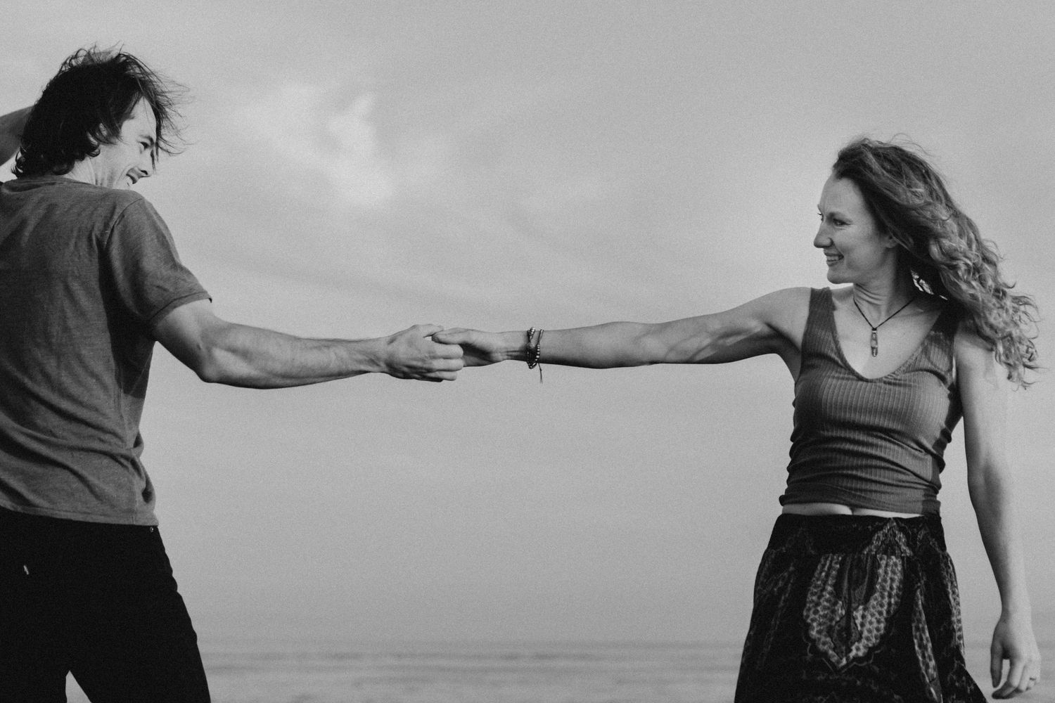 Couple holding hands while standing against ocean backdrop in black and white photo.