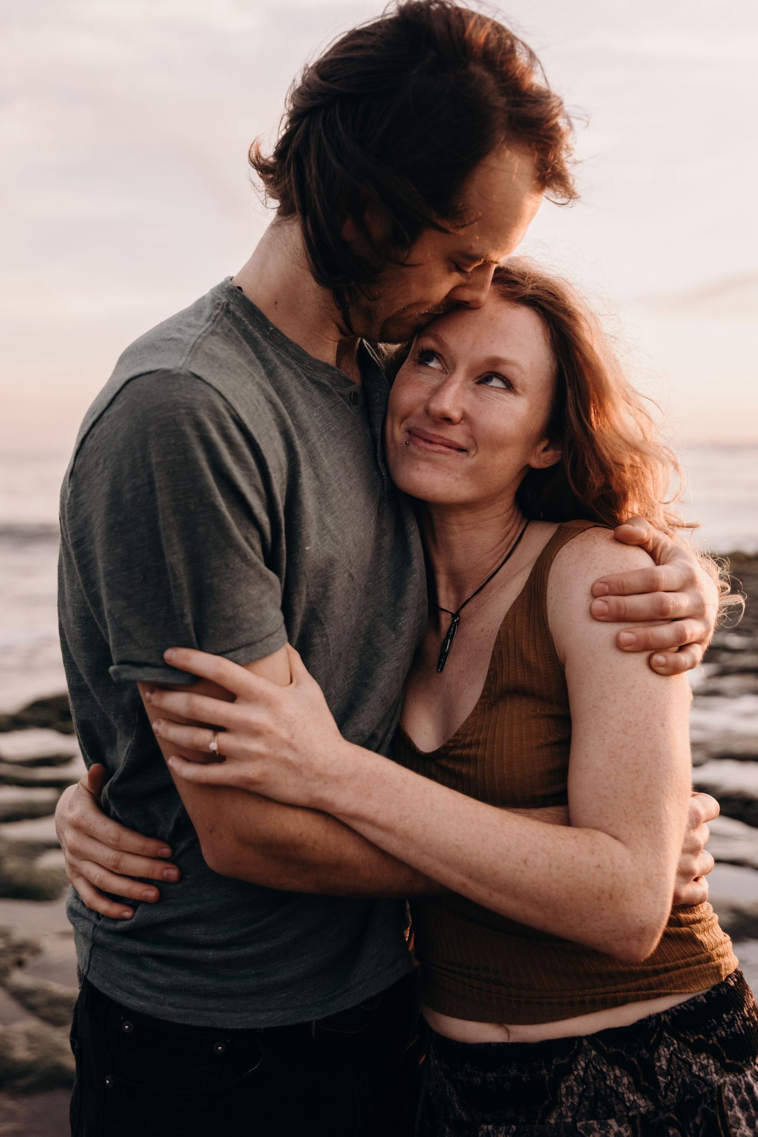 Intimate couple sharing tender moment against rocky coastal backdrop at sunset.