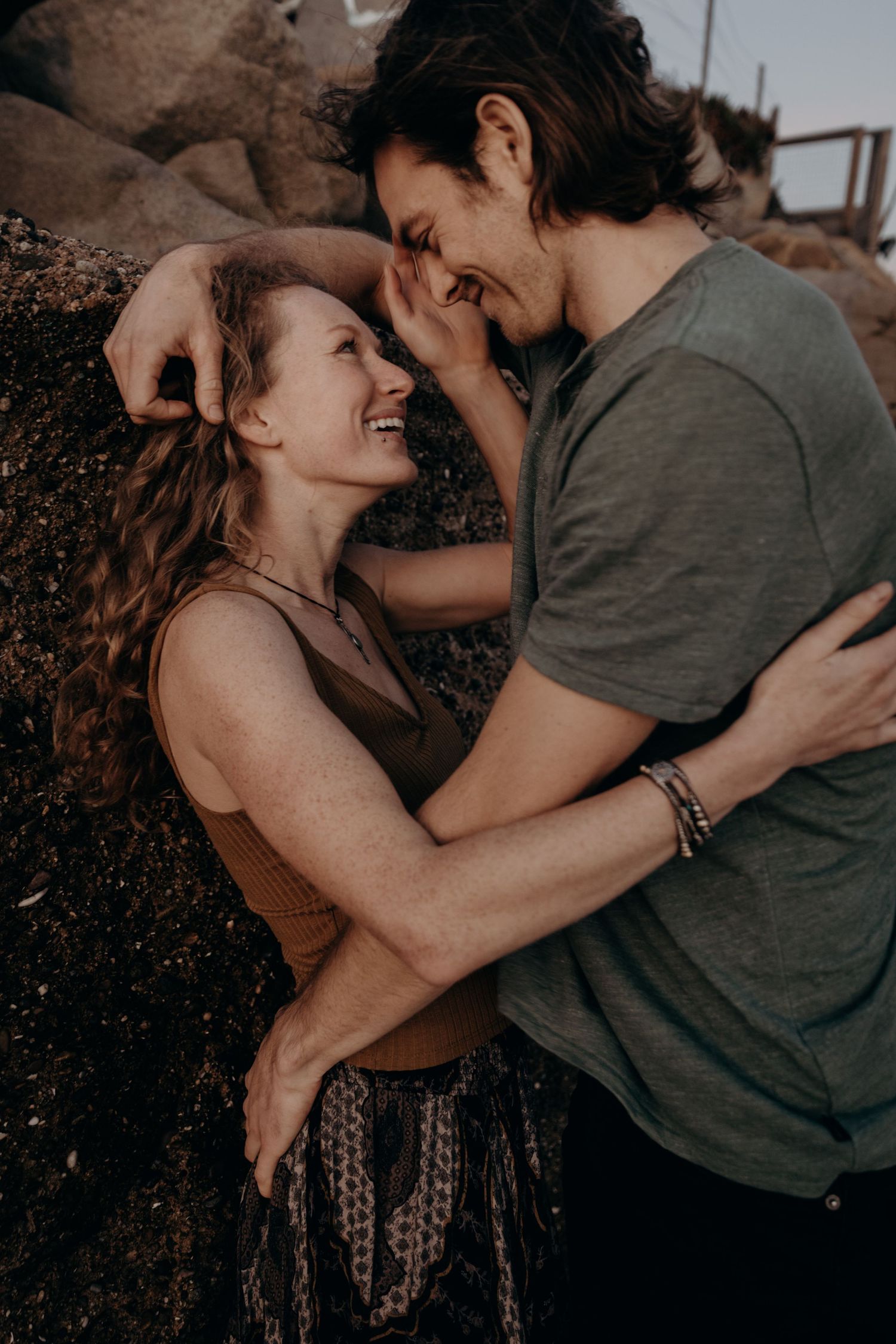 Couple laughing and embracing against rocky wall during golden hour.