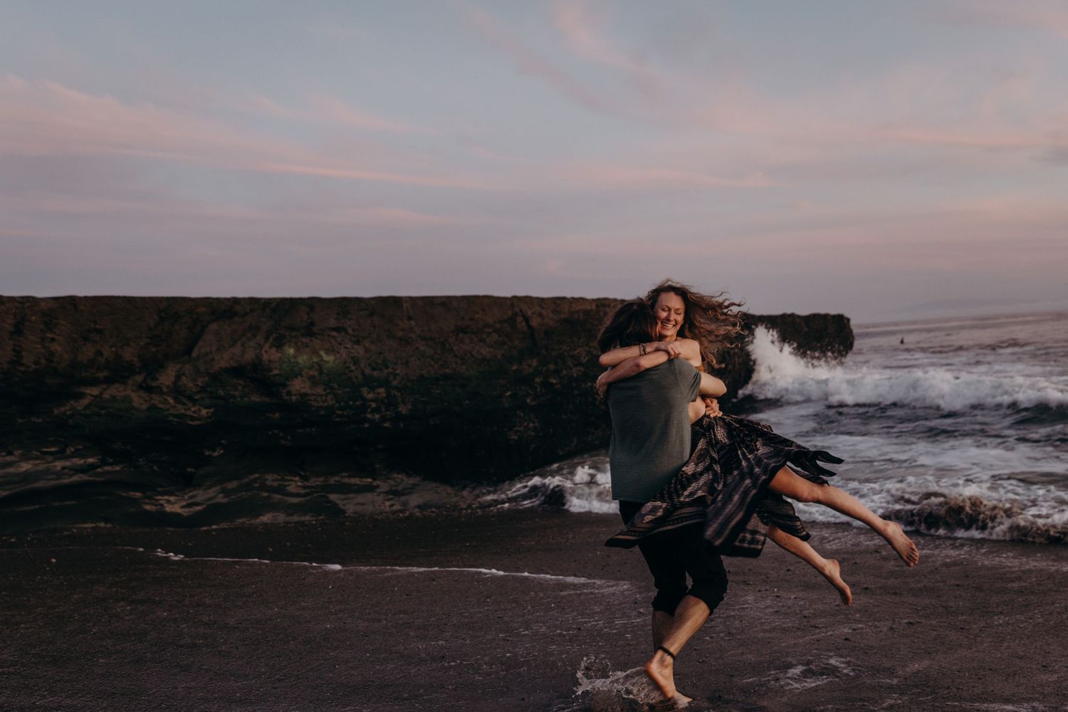 Couple dancing and spinning on rocky beach at sunset.