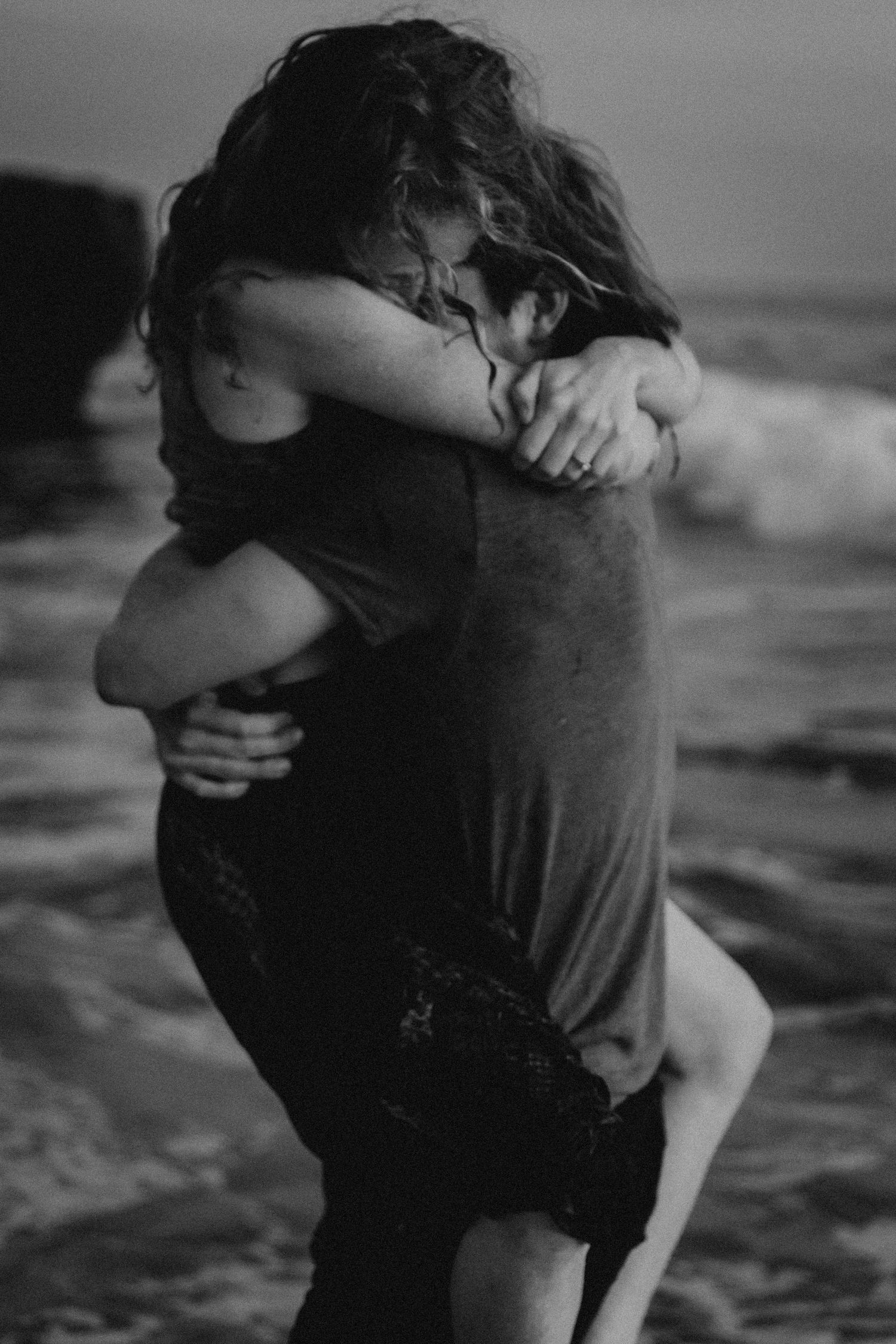 Black and white photo of couple embracing near ocean waves.