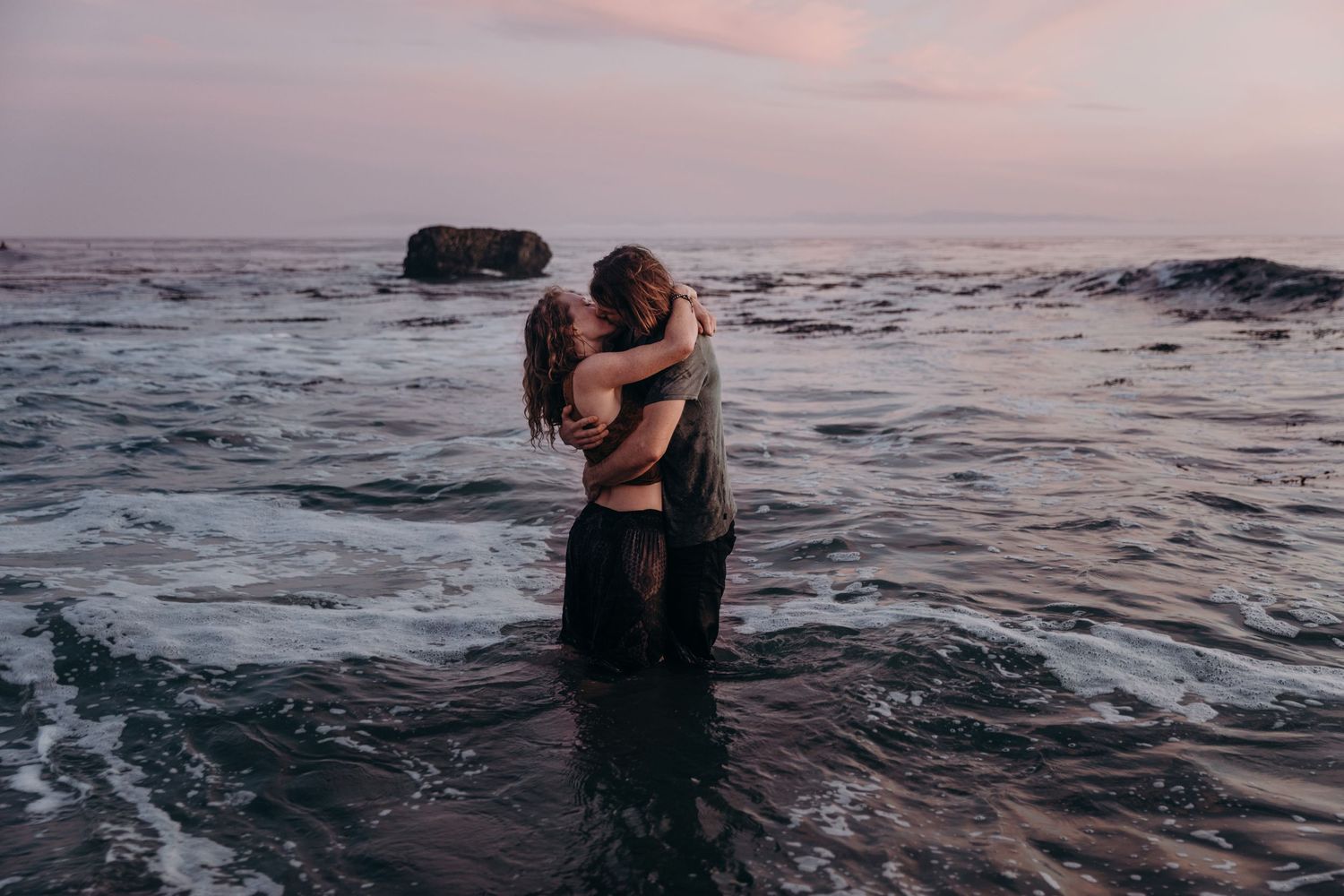 A romantic couple embraces while standing in ocean waves during sunset with rocky coastline in background.