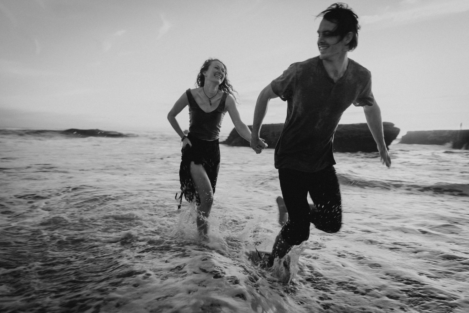 Two people run joyfully through shallow ocean water at the beach in black and white.