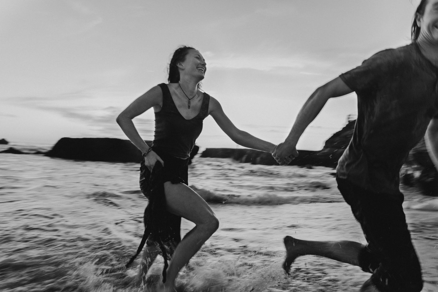 A couple holds hands while running through ocean waves in black and white photography.