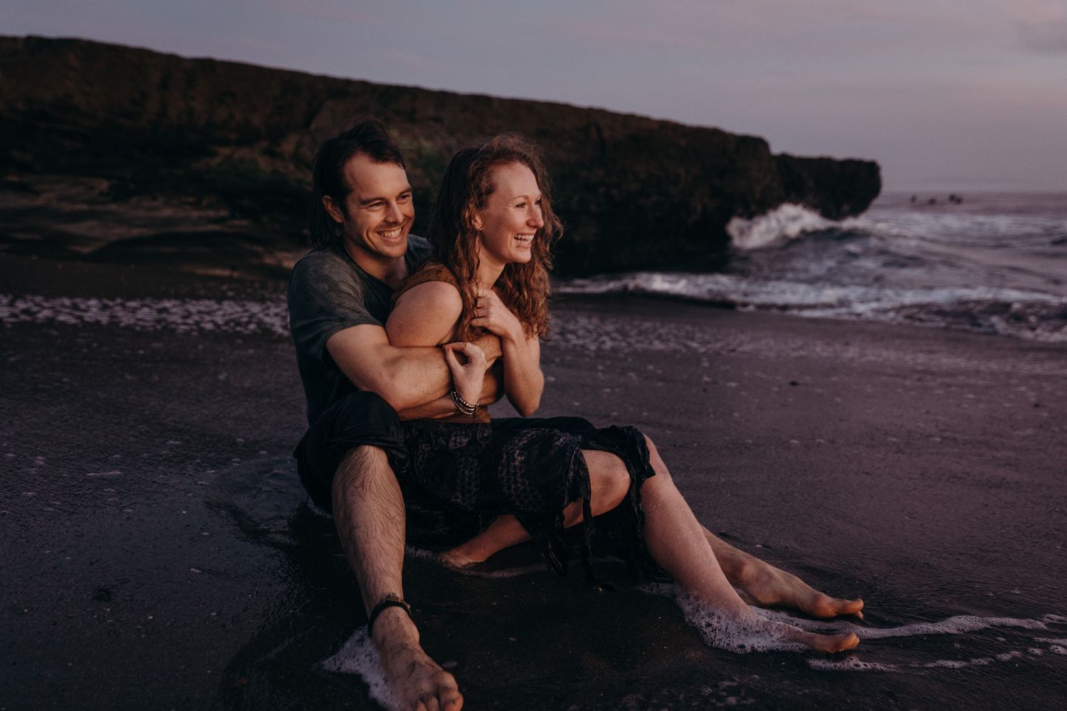A couple sits together on a beach at sunset with rocky cliffs in the background.