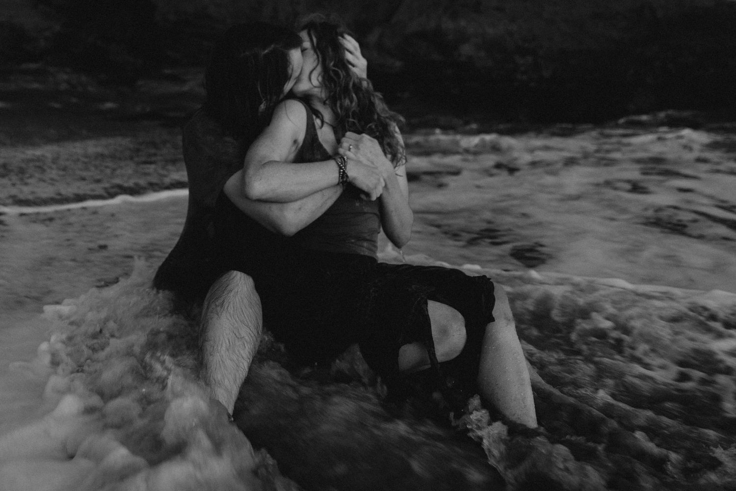 A couple shares an intimate moment while sitting in the ocean surf in monochrome.