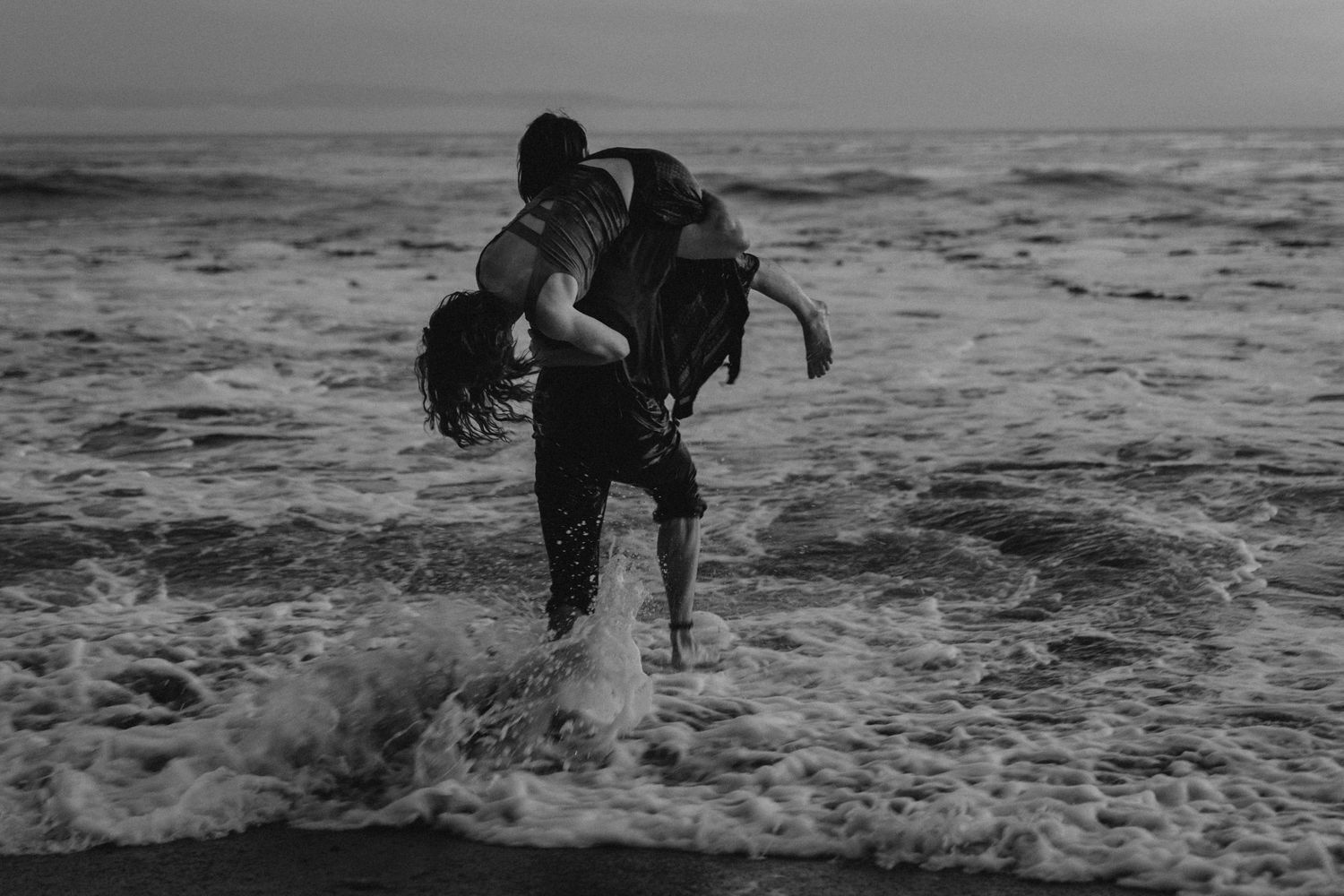 Person carries another through crashing ocean waves in dramatic black and white photography.