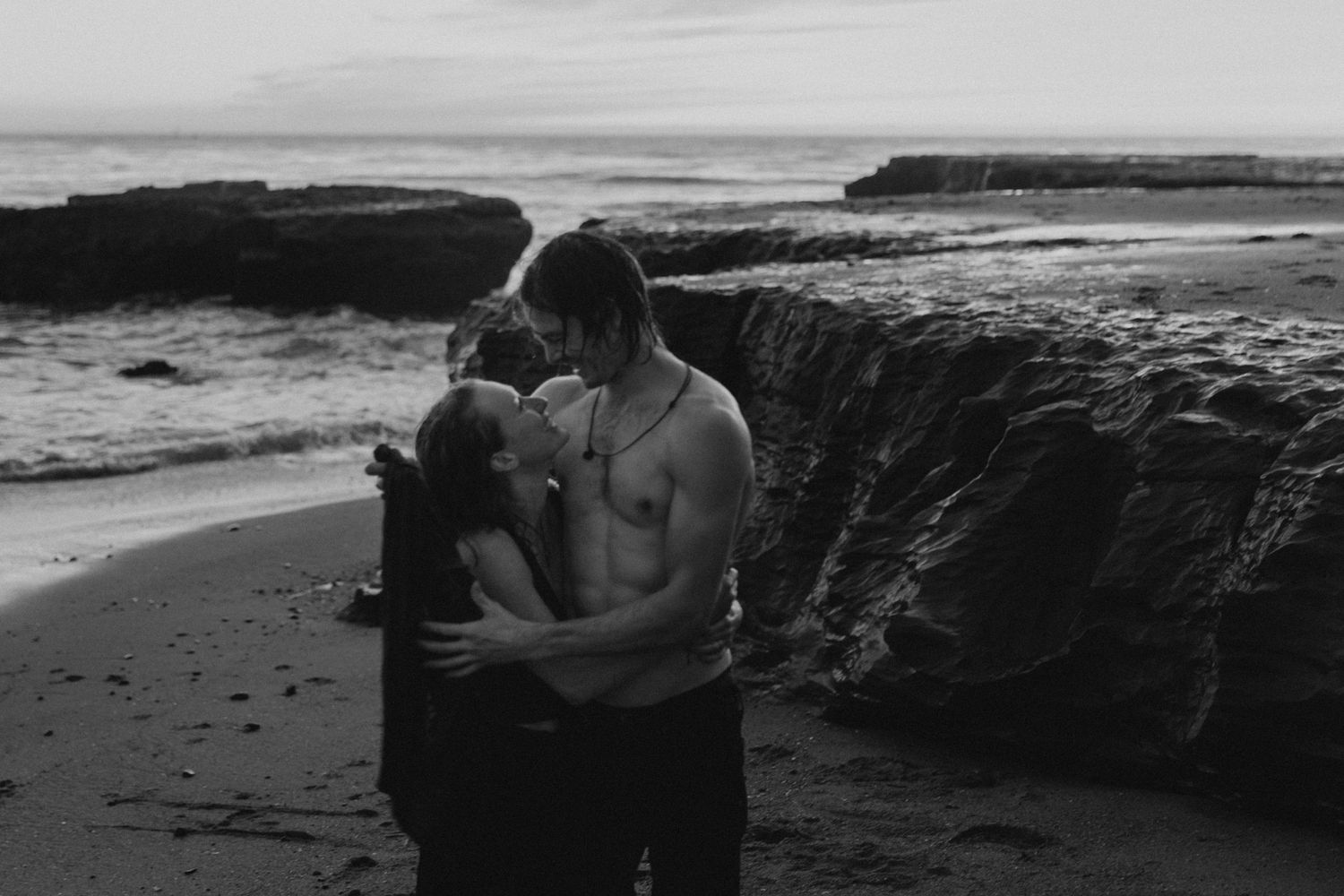 A romantic black and white photo of a couple embracing on a rocky beach at sunset.