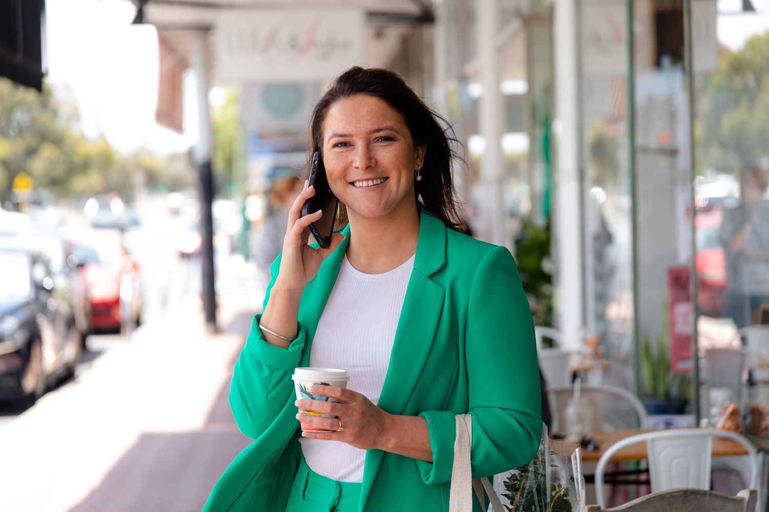 Professional woman in green blazer talks on phone while holding coffee on city sidewalk.