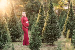 Maternity photo of woman in red dress surrounded by evergreen trees at sunset.