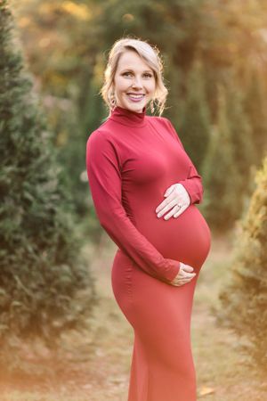 Expectant mother in a form-fitting red dress poses outdoors against an autumn background.