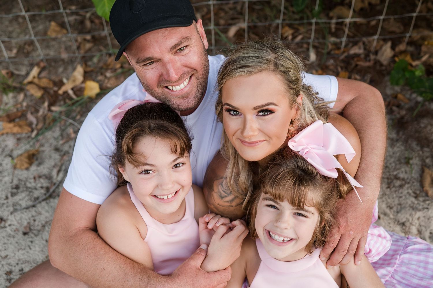 A happy family of four dressed in pink and white outfits poses together outdoors among fallen leaves.