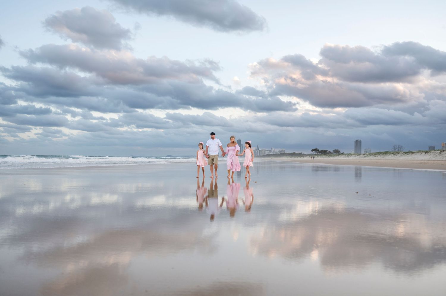A family in coordinated outfits stands on a reflective beach during sunset with dramatic clouds overhead.