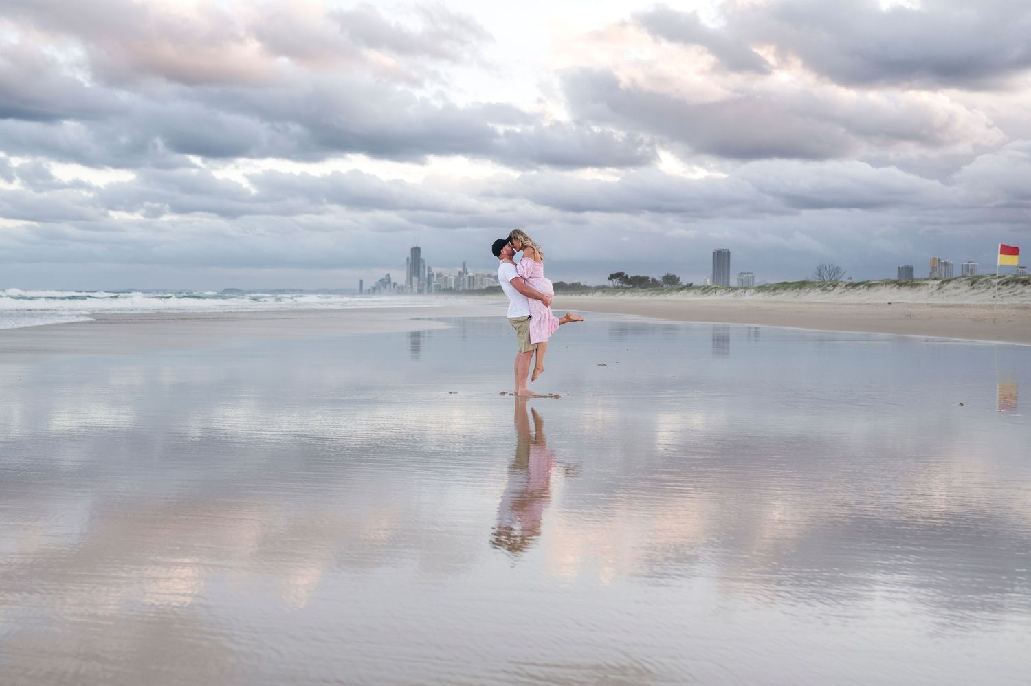 A couple stands on a reflective beach with a cityscape visible in the cloudy background.