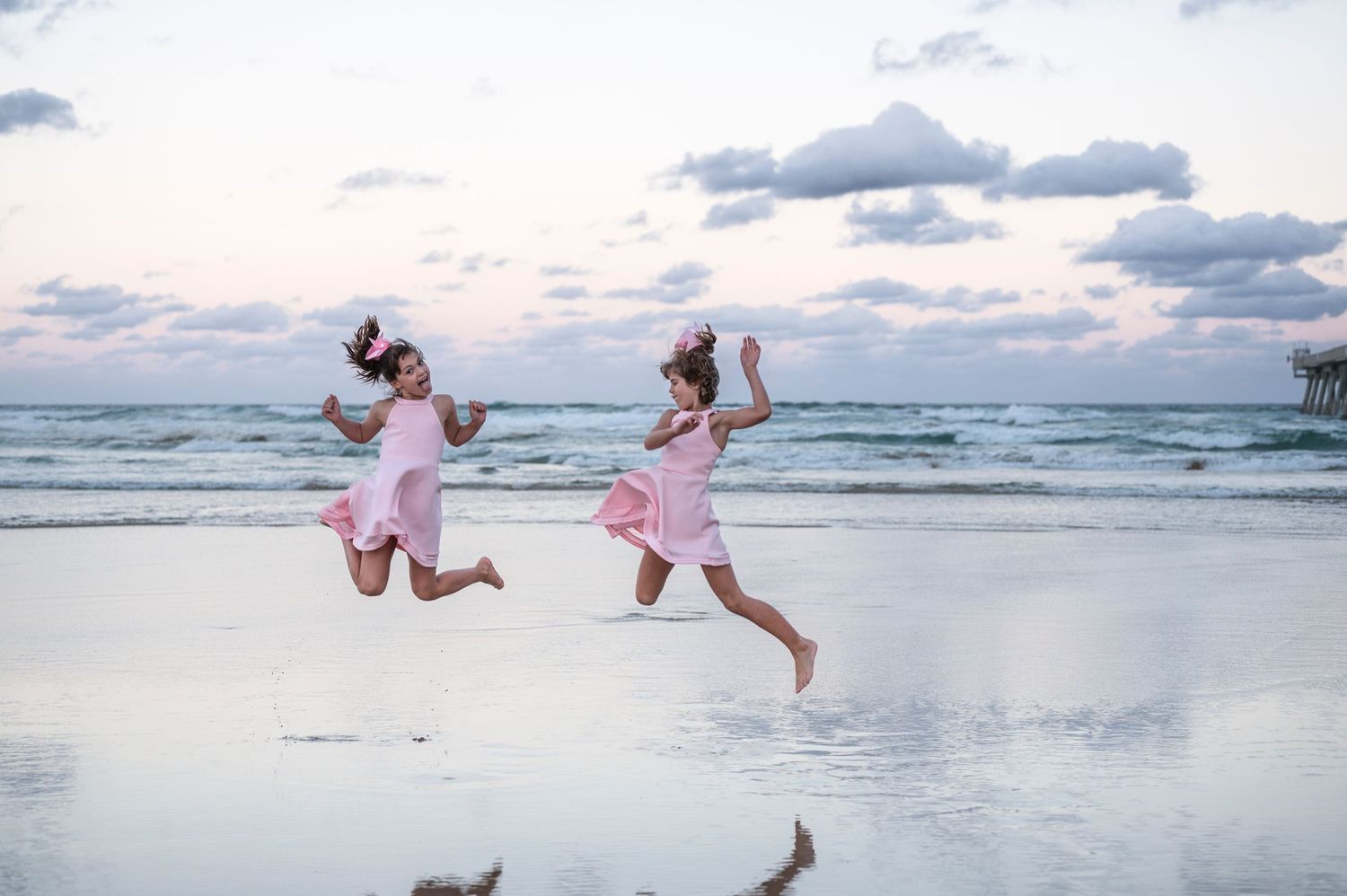 Two children in pink dresses jump playfully on the beach at sunset.