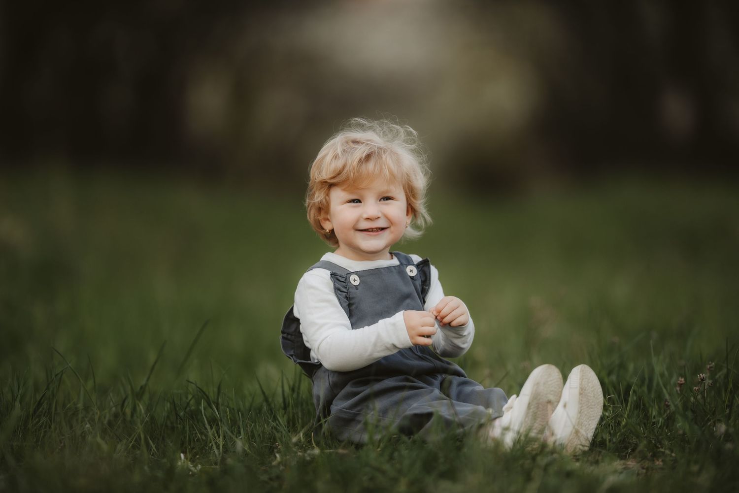 A toddler in overalls sits smiling in green grass during springtime.