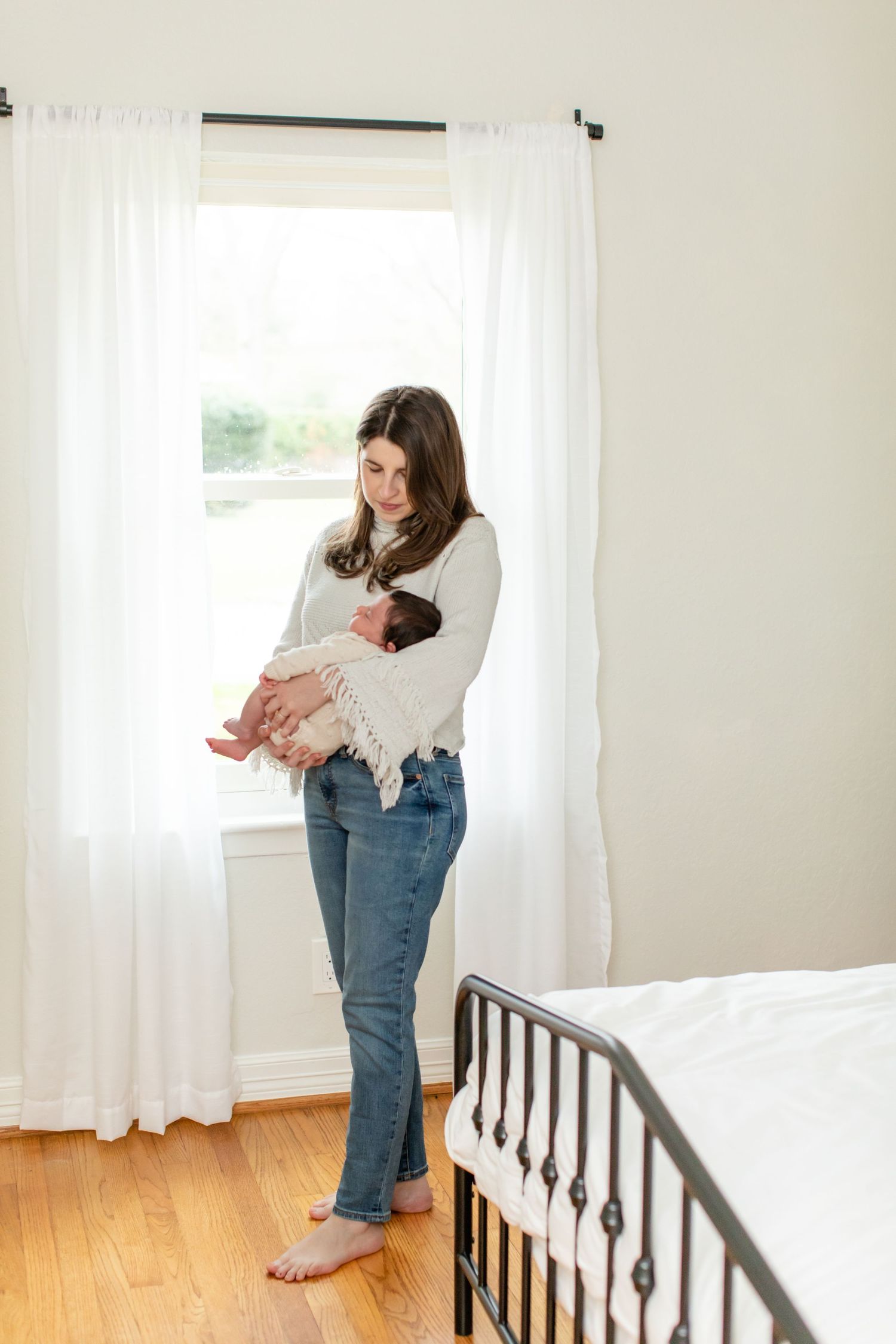 Standing by a bright window, a caregiver cradles a newborn baby in a white nursery.