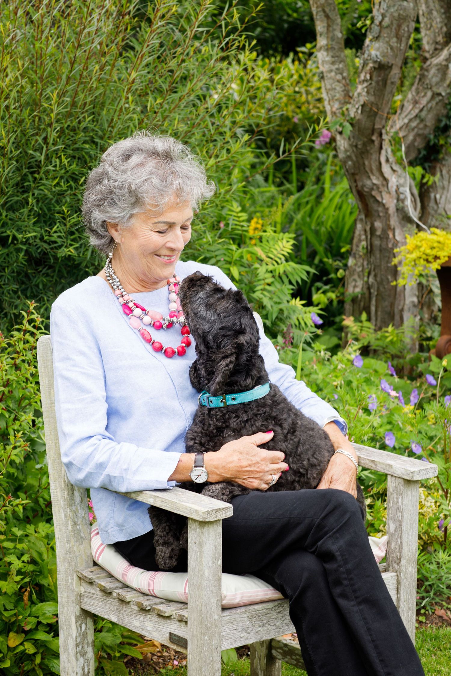 Person sitting in wooden garden chair with small black dog in beautiful garden setting.