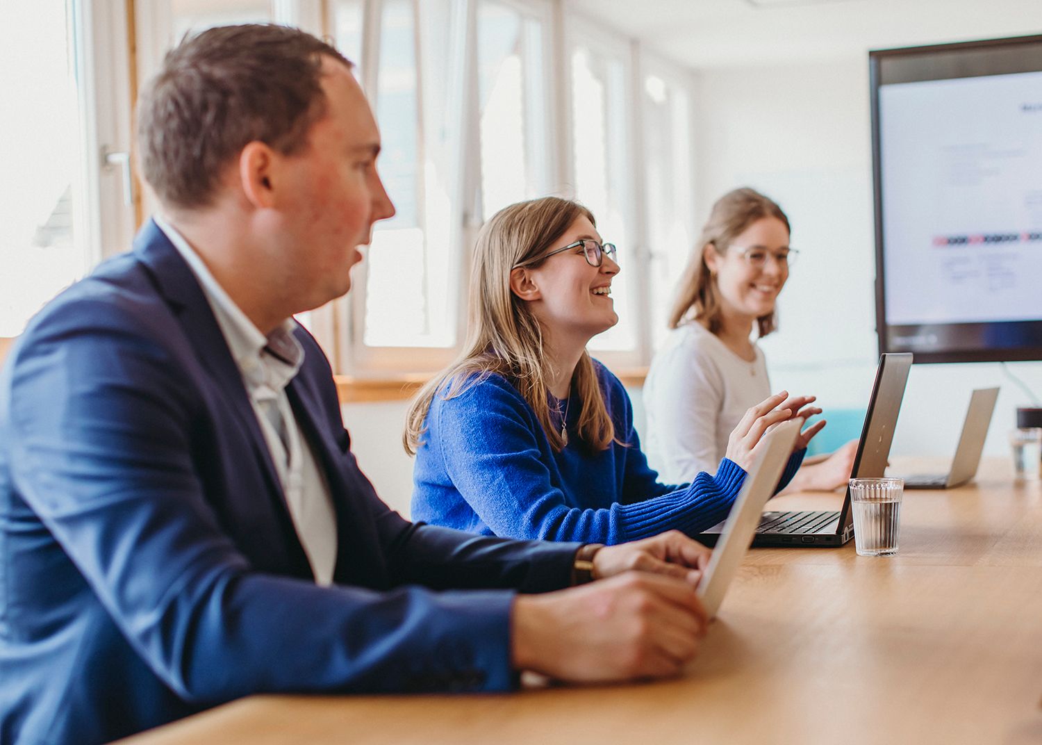 Team members working together at conference table with laptops in bright office space.