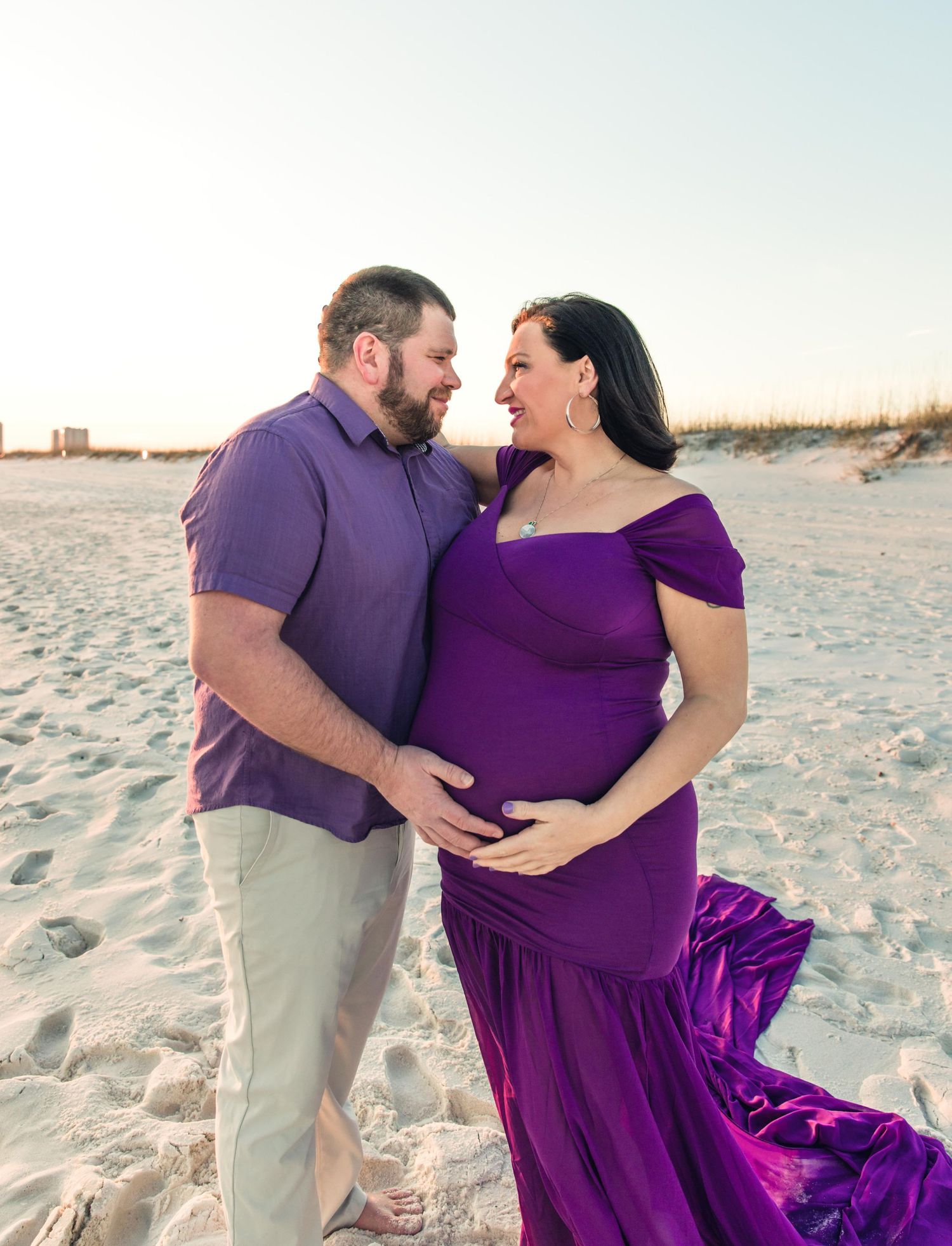 Maternity beach portrait with couple in coordinating purple outfits on sandy shore.