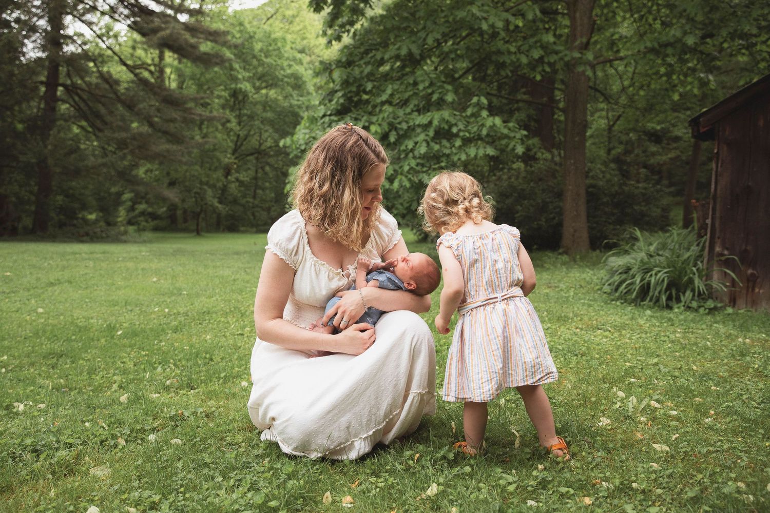 Outdoor Photoshoot of a mother in a dress holding a newborn with two year old sister kissing the babies head.