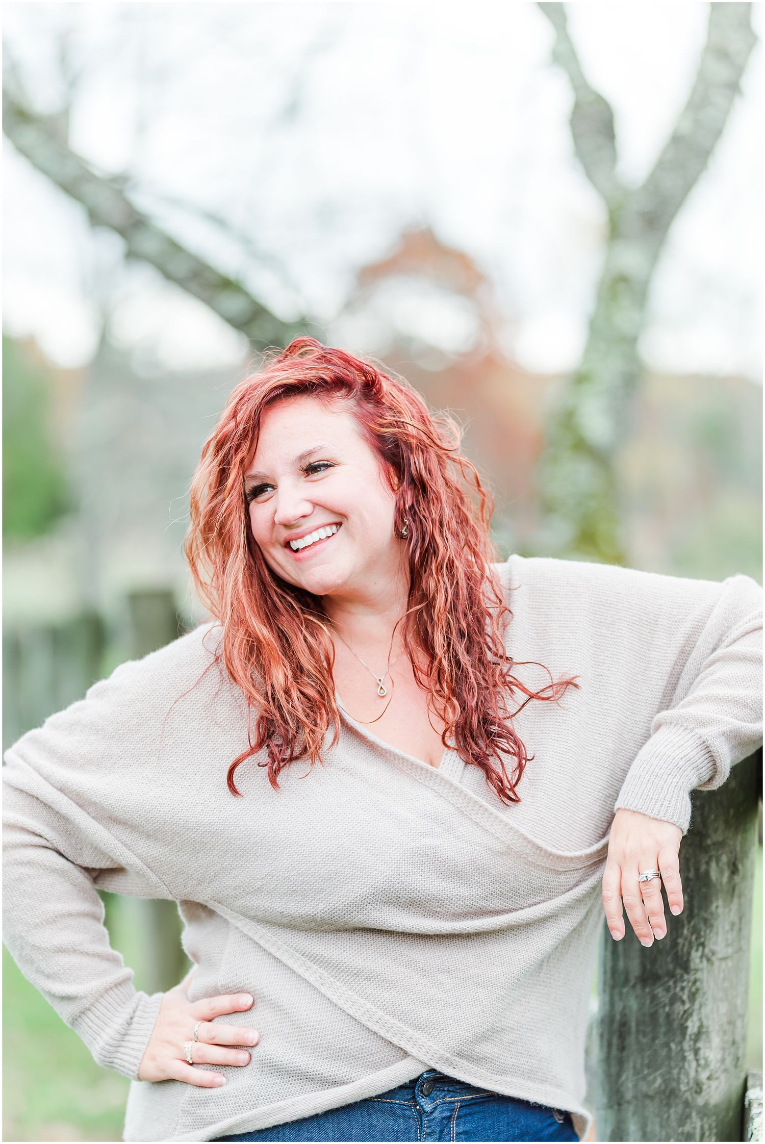 Person in a gray sweater smiles while leaning against a tree in an outdoor portrait setting.
