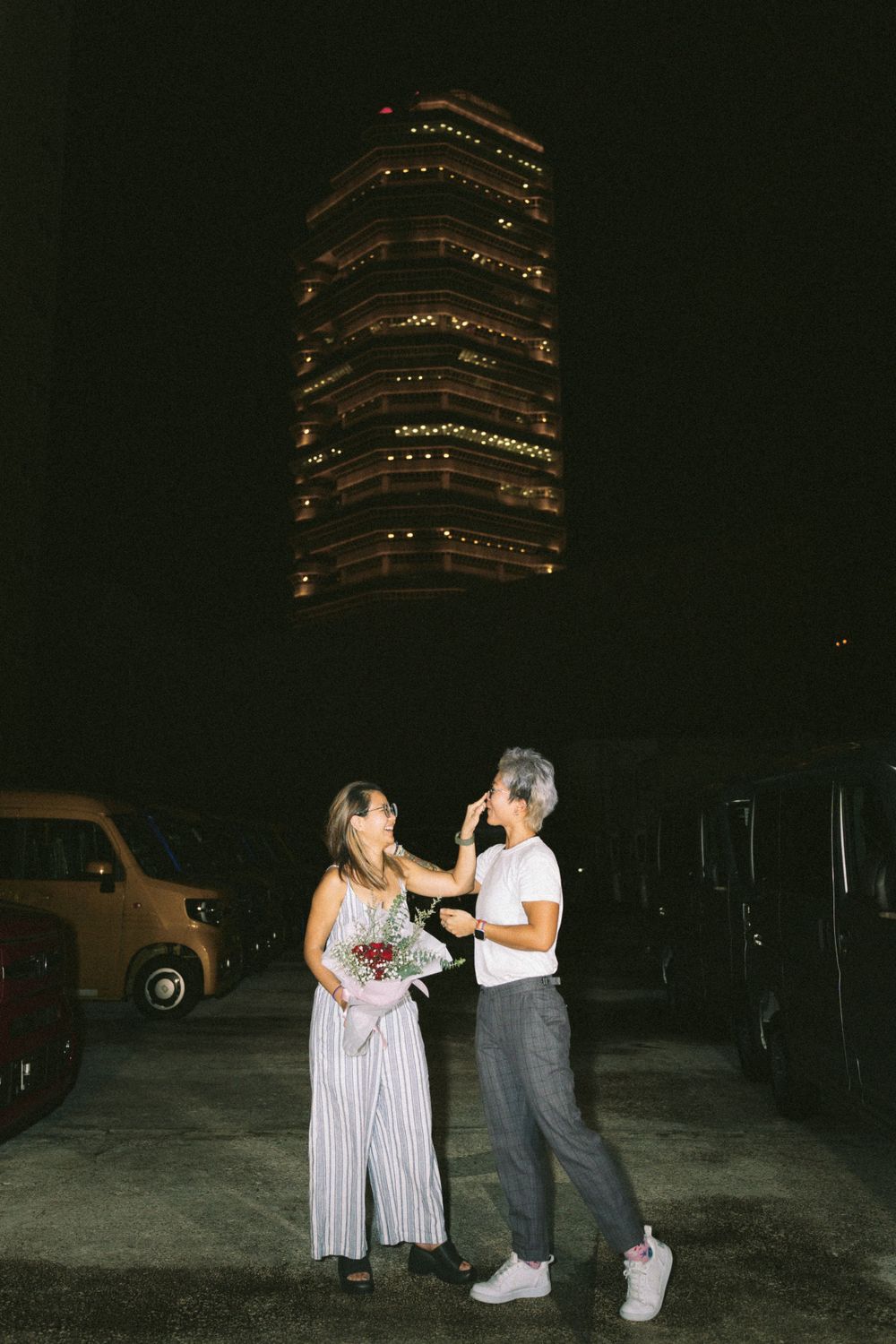 A couple sharing a moment in front of an illuminated high-rise building at night.