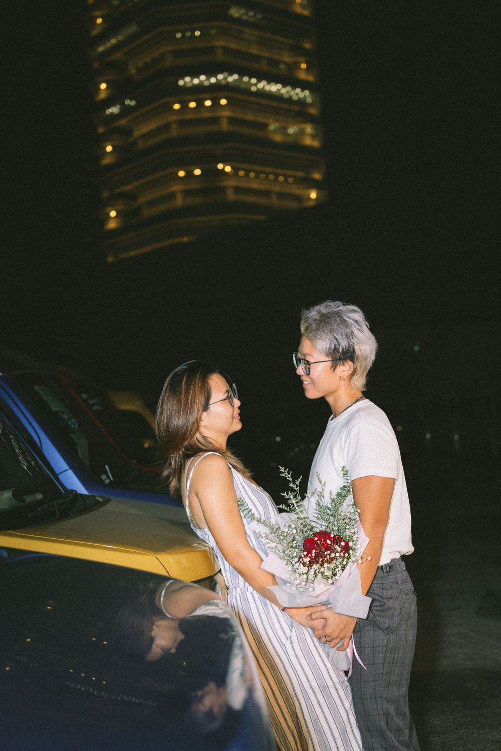 Two people embracing while holding flowers in front of a car with a glowing building behind them.
