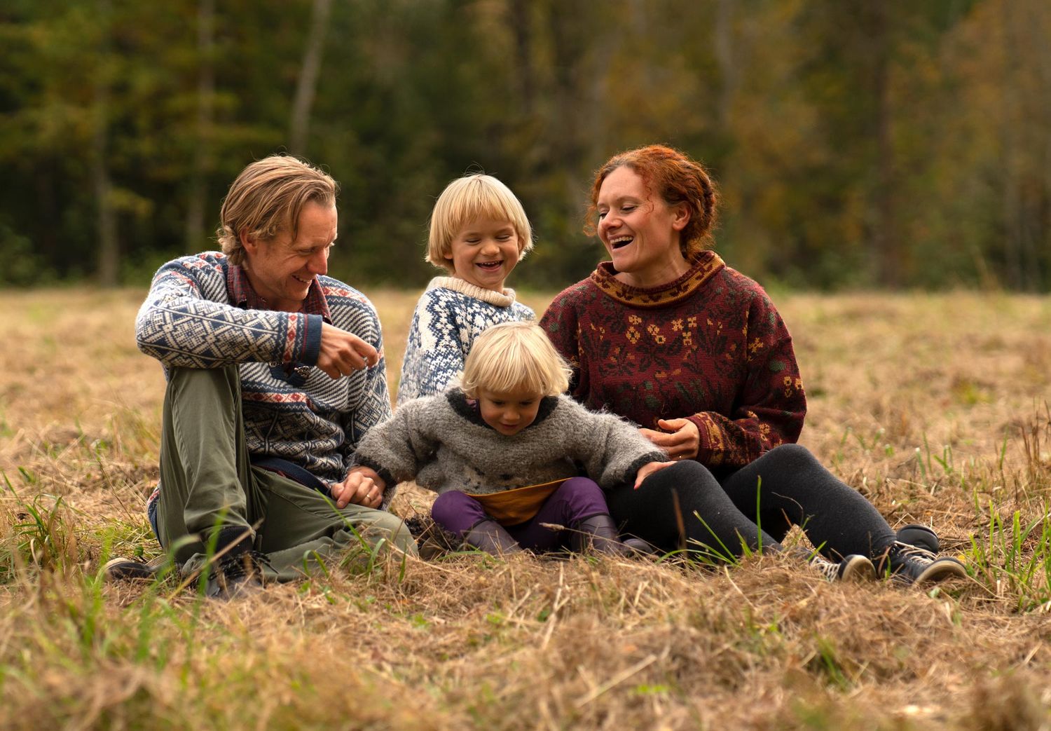 Mamma, pappa och två barn sitter utomhus i gräset en höstdag med skogen i bakgrunden