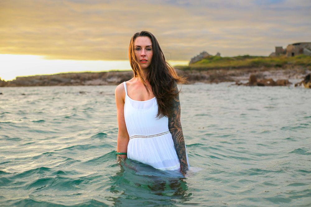 A person in a white dress stands in ocean water at sunset with rocky coastline in background.