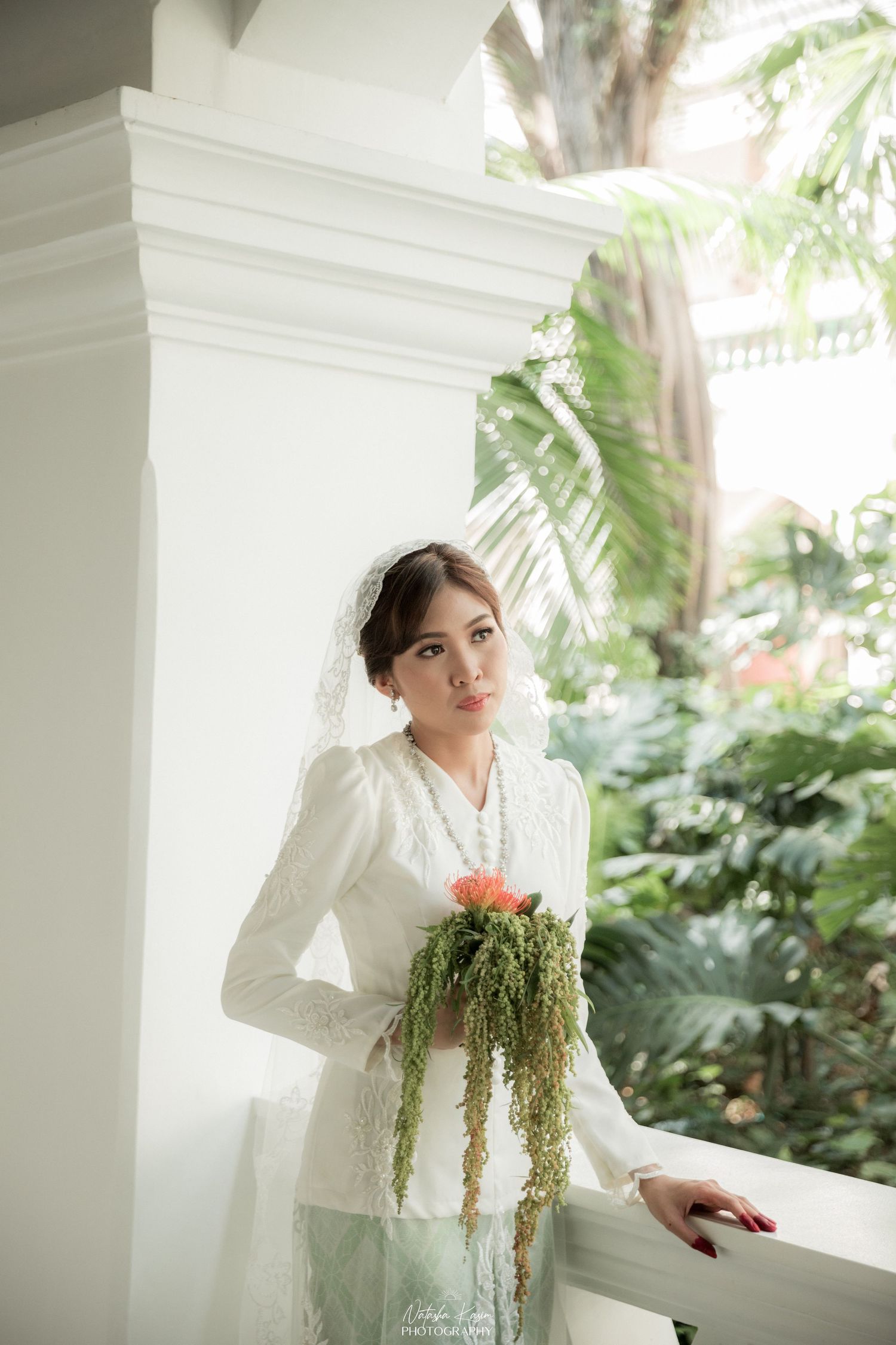 A person in white attire poses elegantly against a white colonial pillar surrounded by lush tropical foliage.